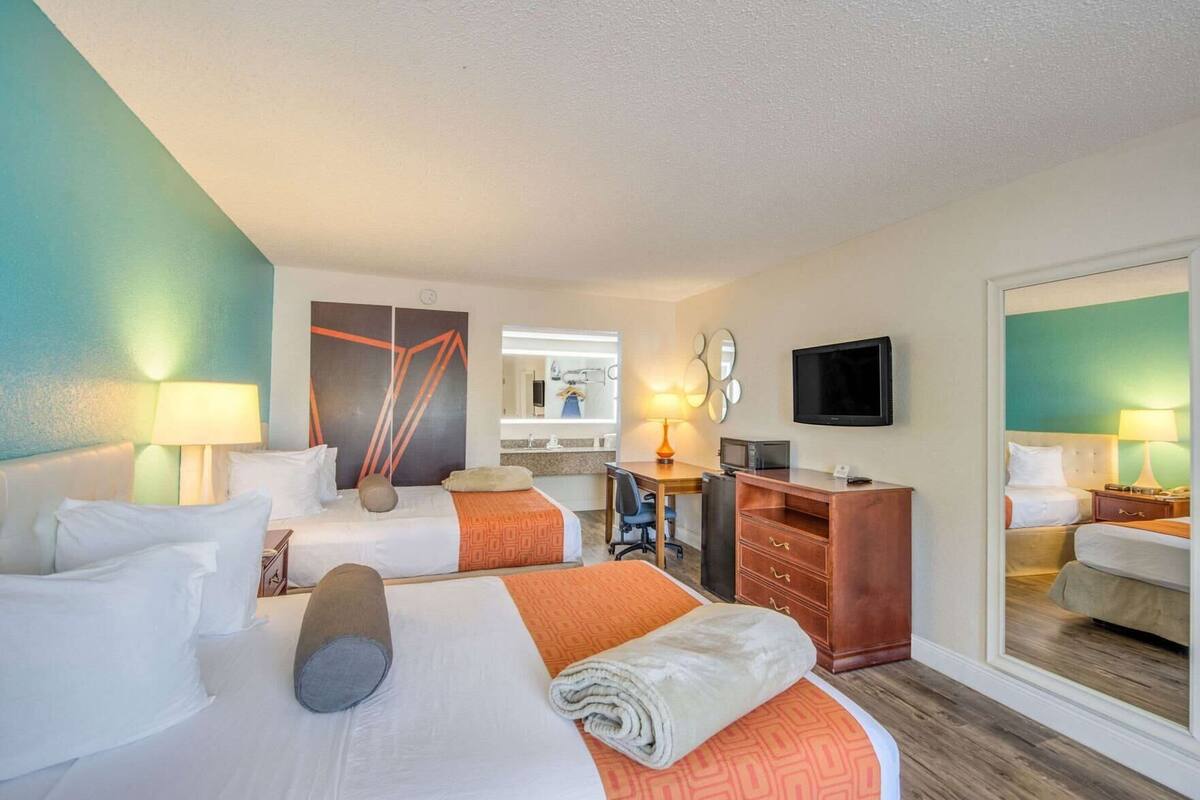 A hotel room with two beds, a desk, a TV, and a bathroom visible in the background.