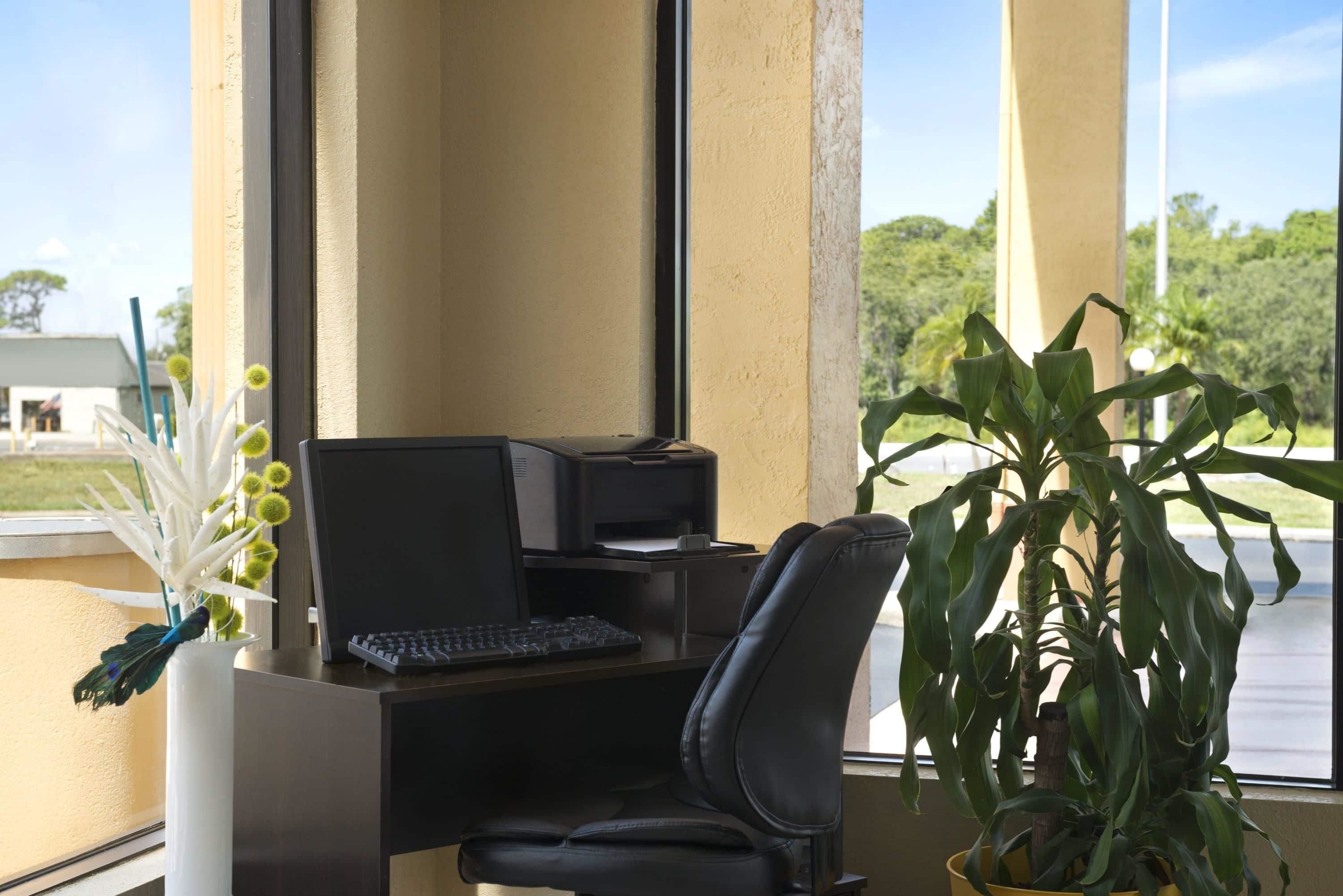 Un escritorio con computadora, teclado e impresora, una silla de oficina negra y una planta en maceta.