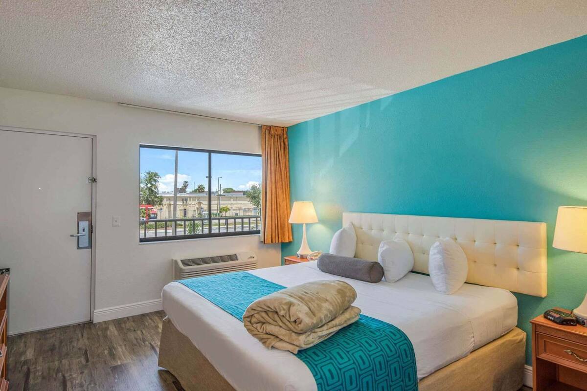 A hotel room with a large bed, a window with a view, a door, and a small table with a lamp.