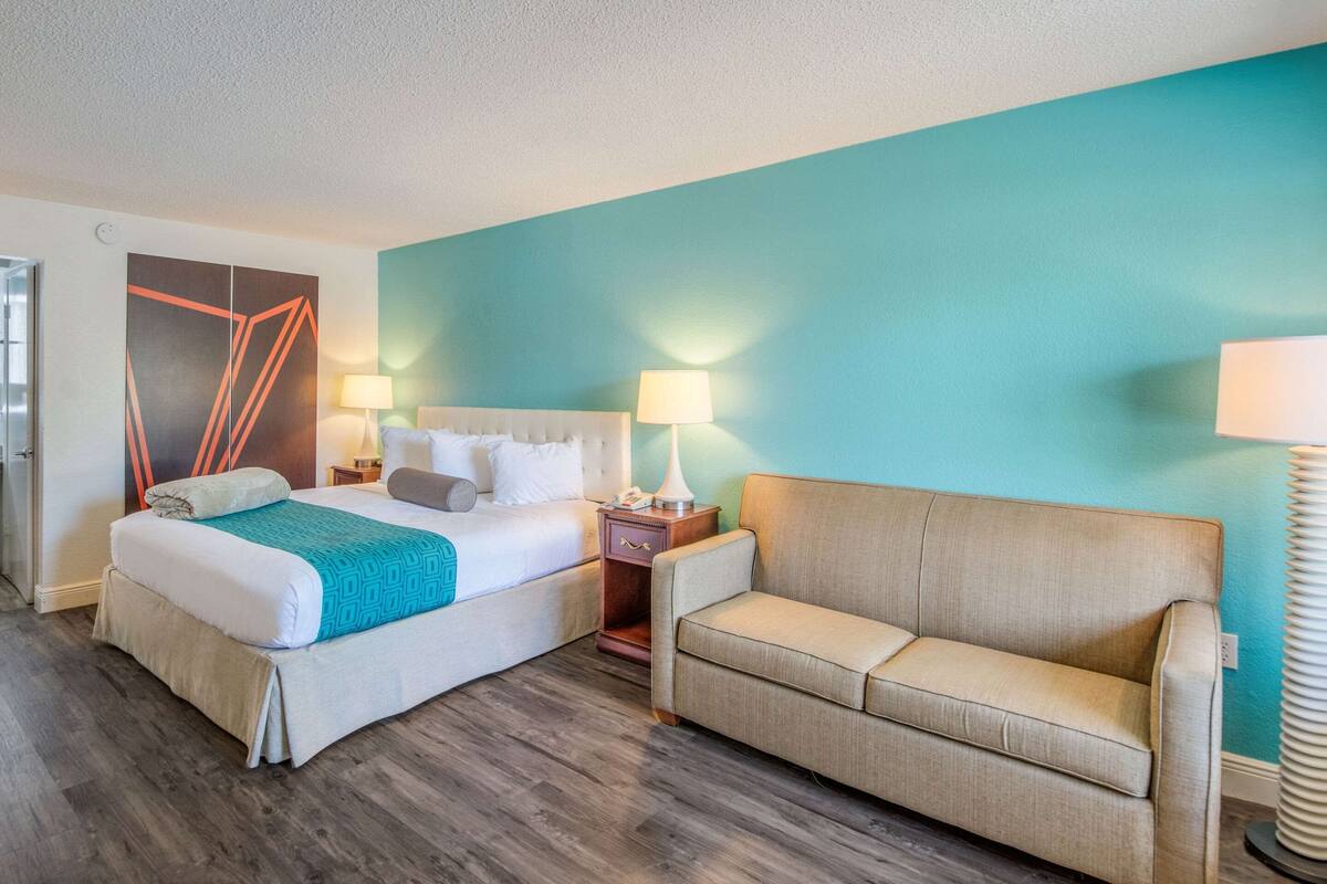 A hotel room with a bed, a sofa, a nightstand with a lamp, and a wall with geometric art.