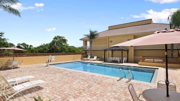 Una piscina con tumbonas y sombrillas, rodeada por un patio de ladrillo y un edificio con techo de tejas.