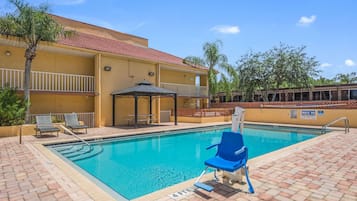 A swimming pool with lounge chairs, a covered seating area, and a building with a red-tiled roof.