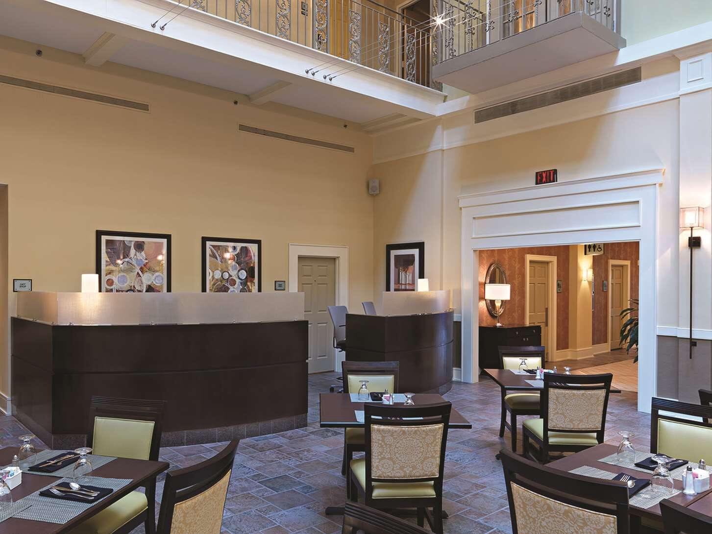 A hotel lobby with a reception desk, multiple dining tables, and chairs. There are framed artworks on the walls, a staircase, and an exit sign.