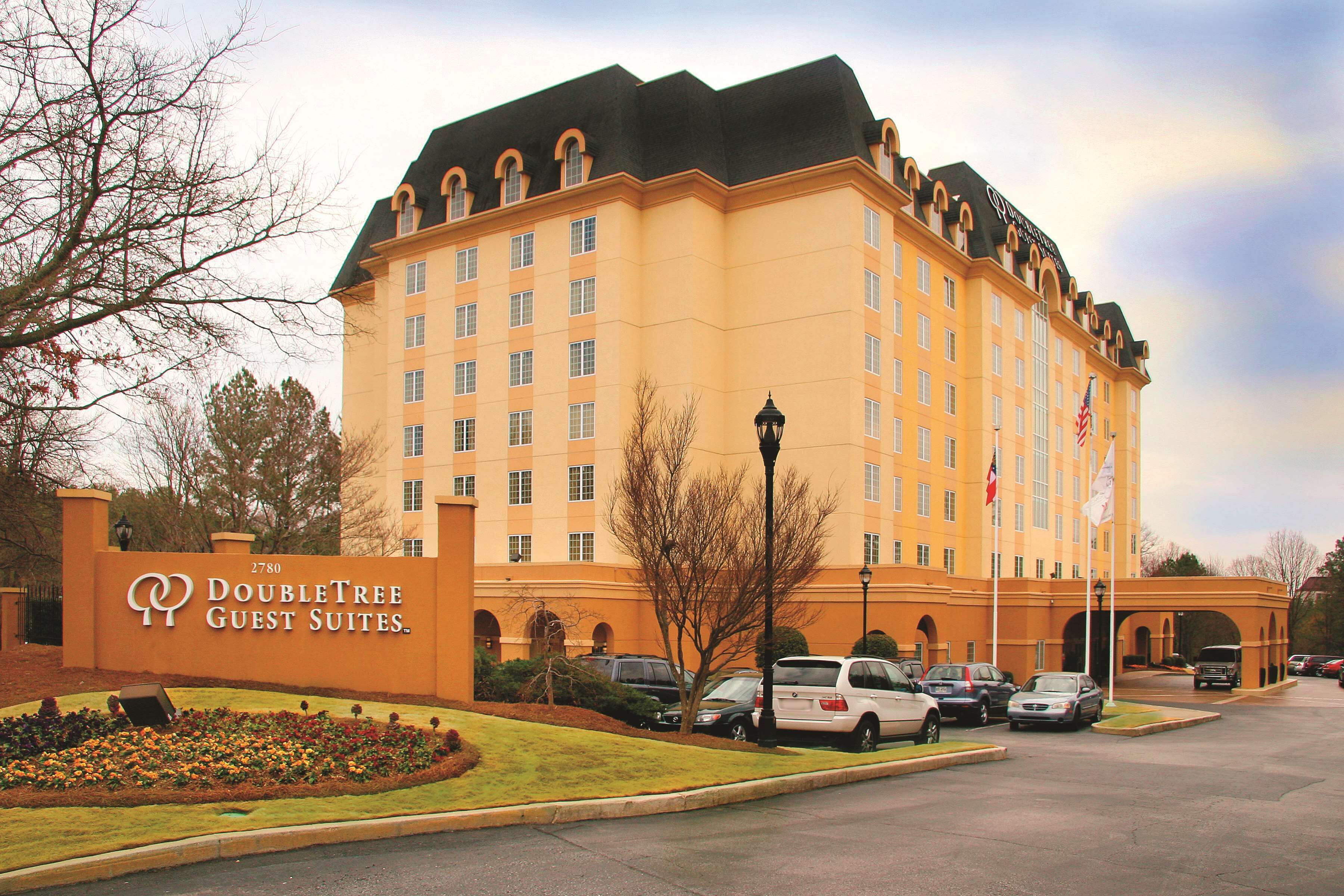 A large hotel with a sign reading "DoubleTree Guest Suites." It has multiple floors, a parking lot with several cars, and a well-maintained lawn area.