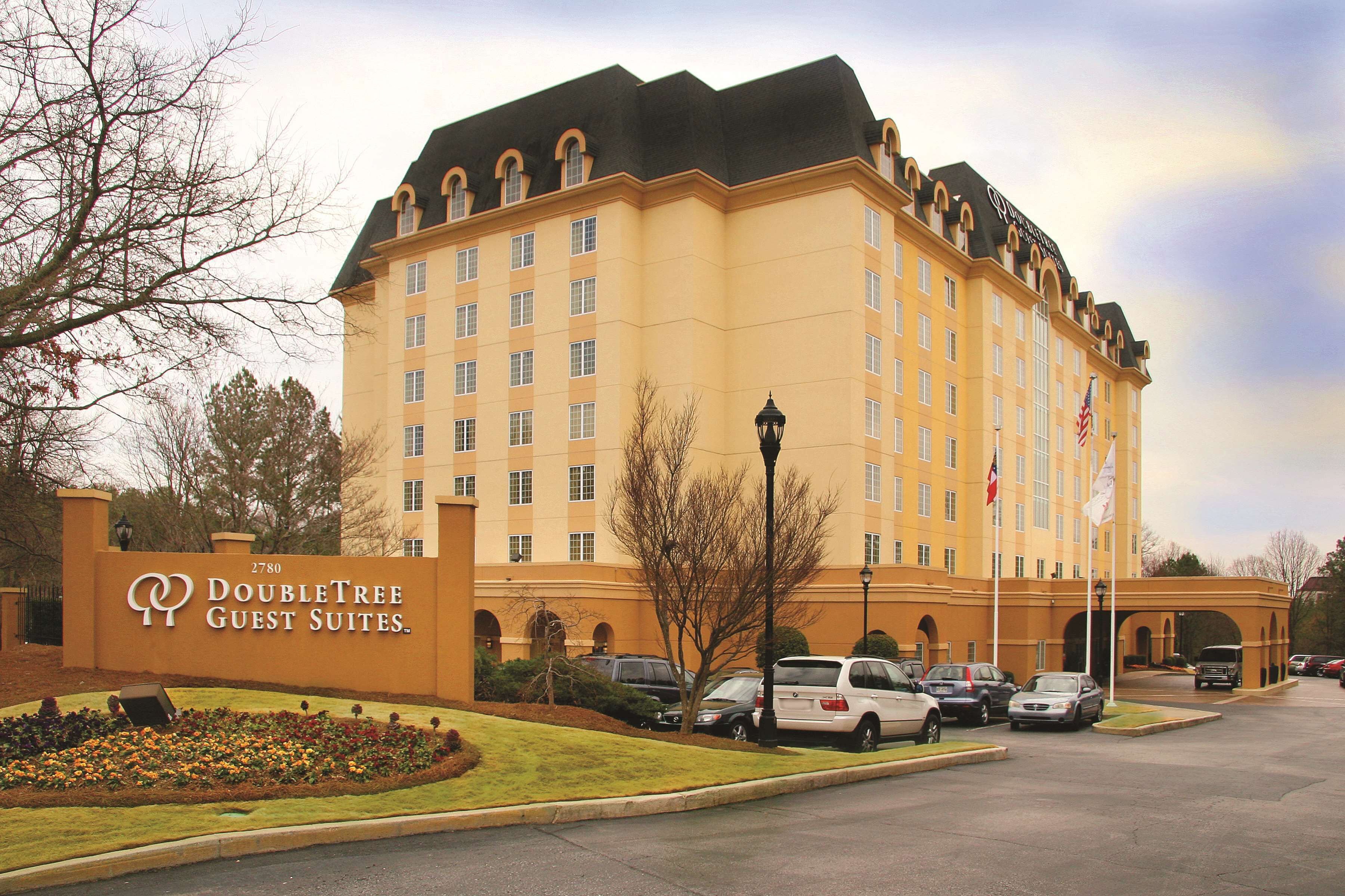 A large hotel with a sign reading "DoubleTree Guest Suites." It features a parking lot with several cars, a well-maintained lawn, and a flagpole with flags.