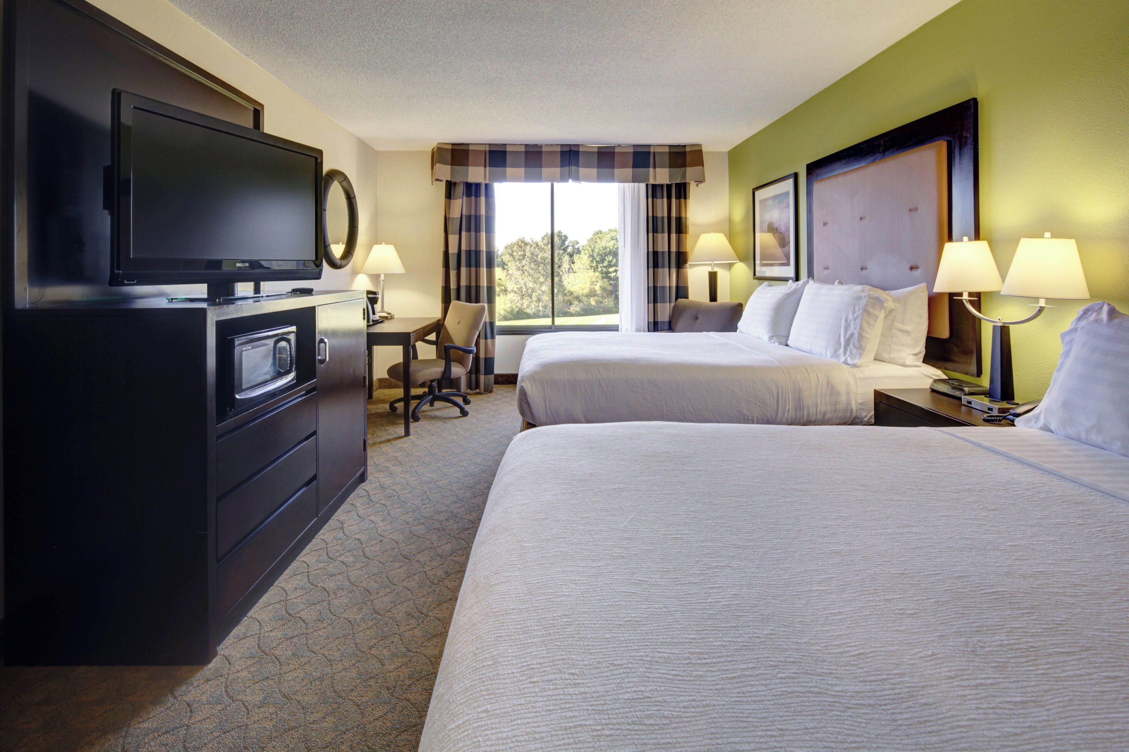 A hotel room with a television, a desk, two beds, and a window with curtains.