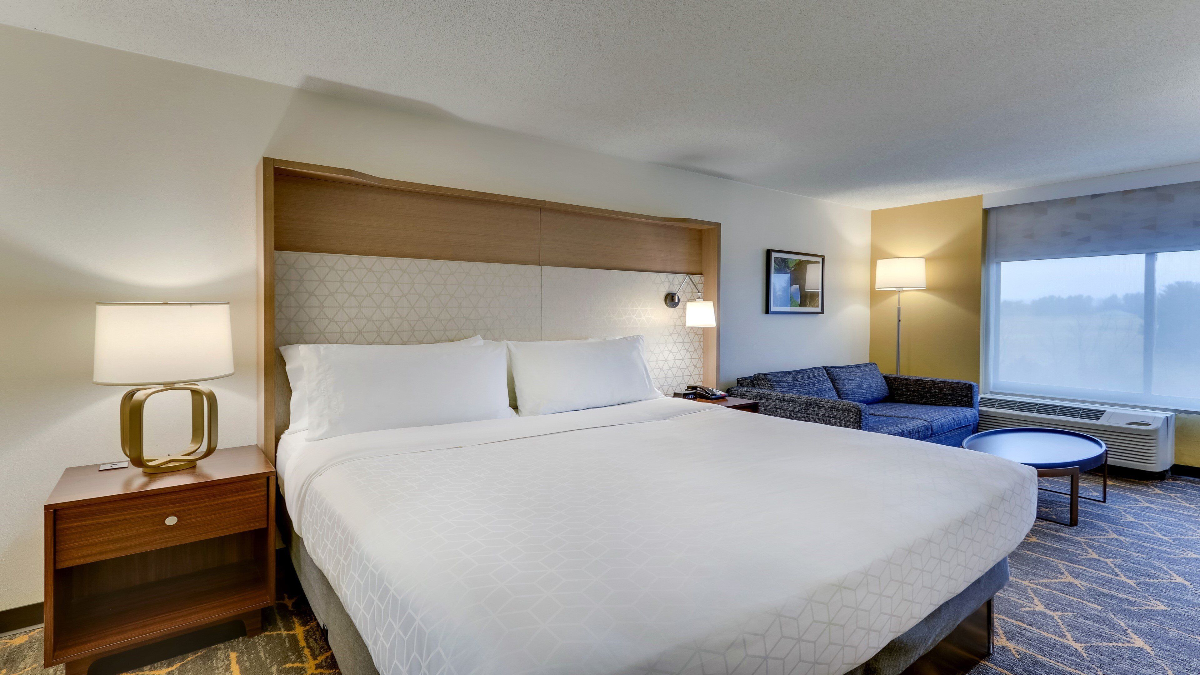 A hotel room with a large bed, a bedside table with a lamp, a sofa, a small table, and a window with a view.