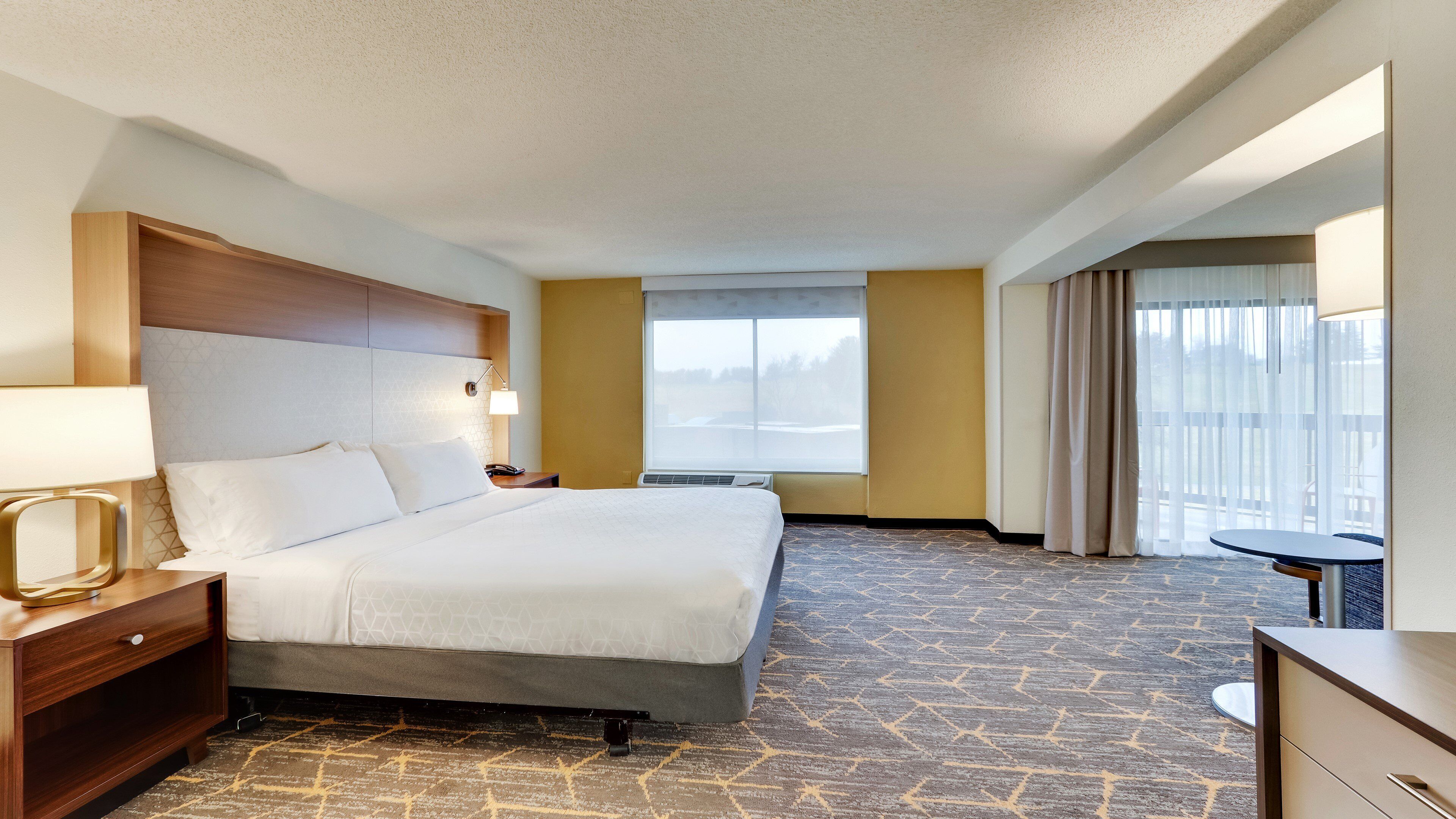 Habitación de hotel con cama, mesita de noche, lámpara, ventana y vistas al exterior.