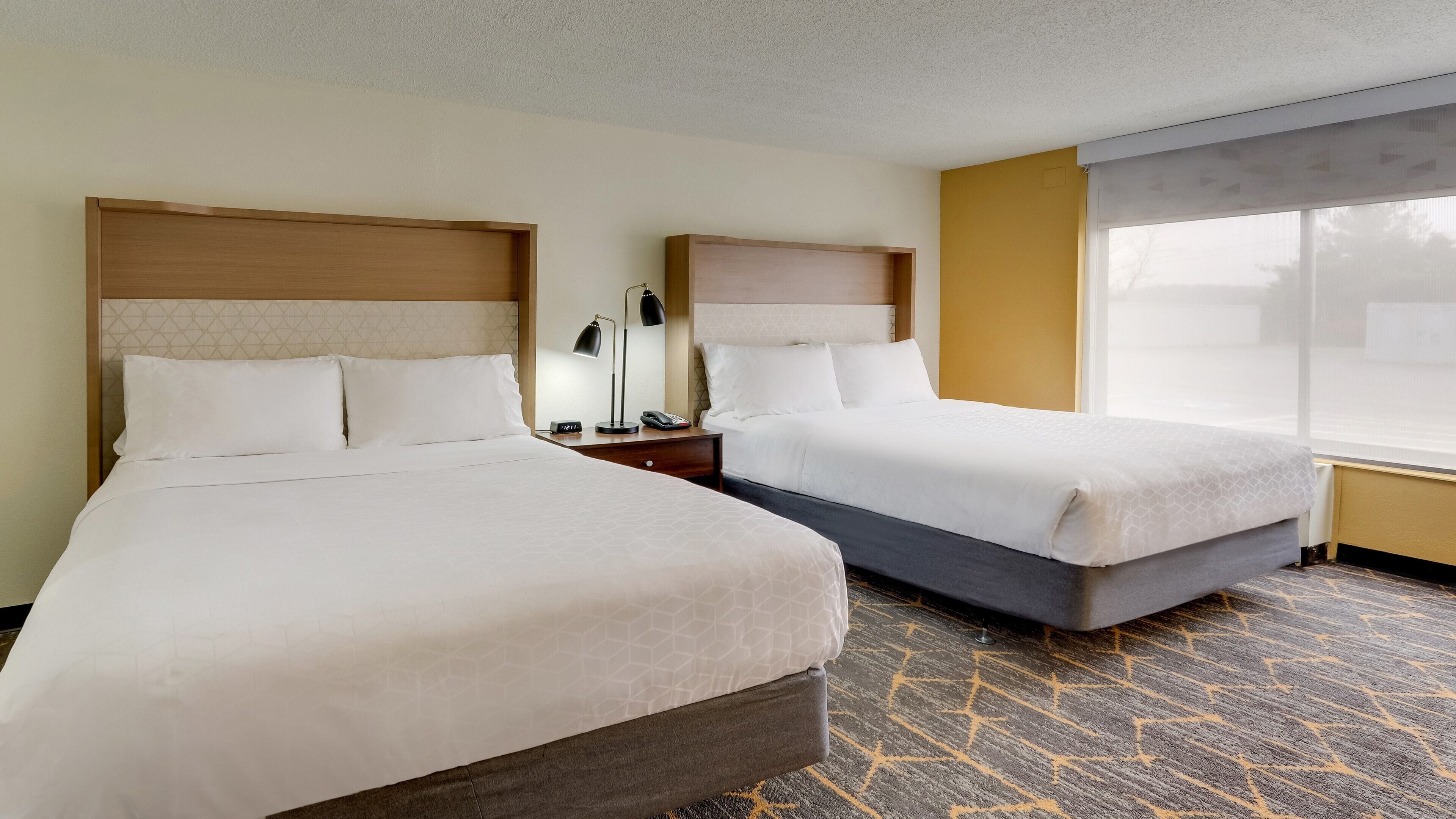 A hotel room with two beds, a nightstand with a lamp, and a large window.