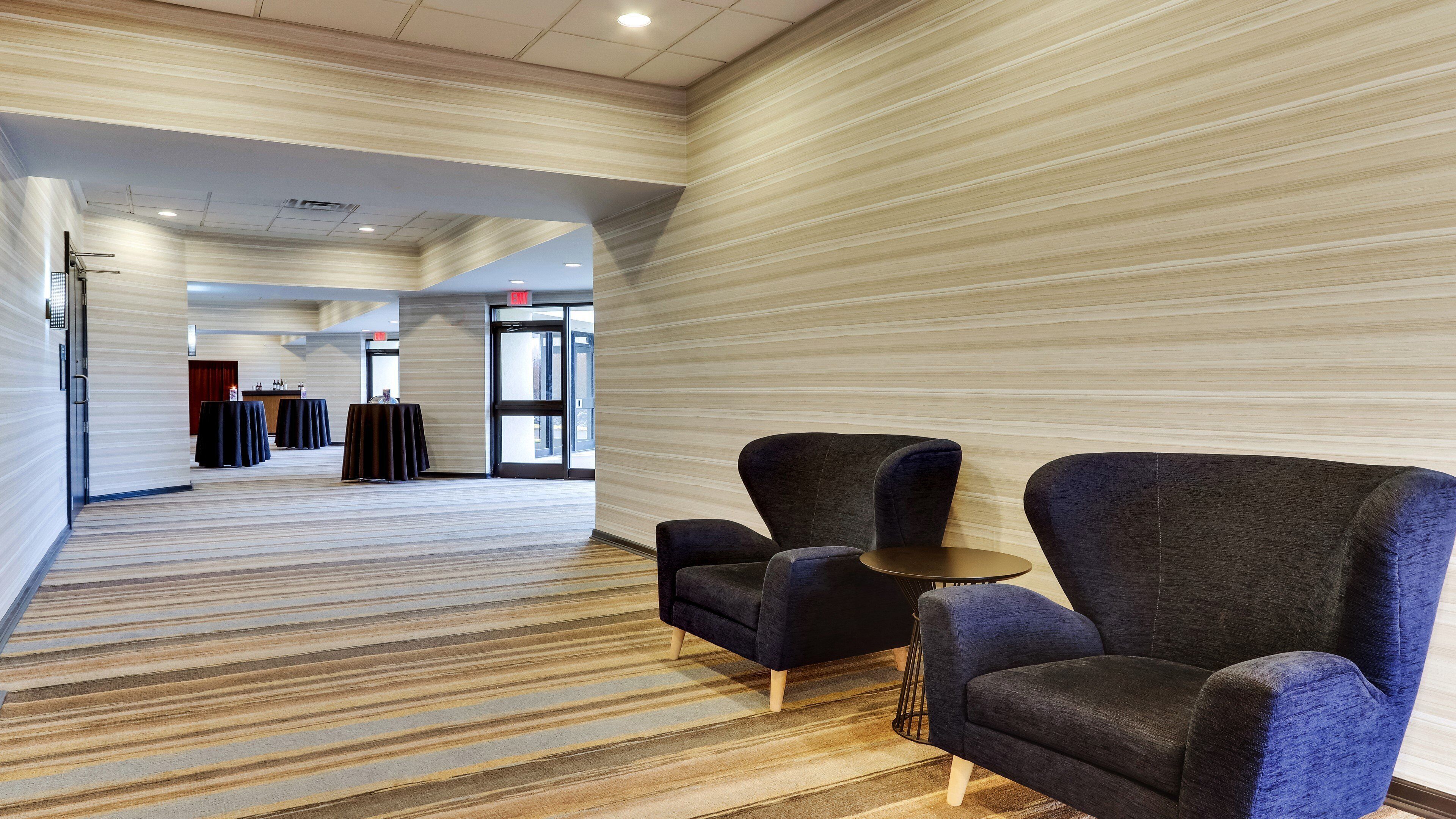 A hotel lobby with striped carpet, two armchairs, a small table, and a reception desk in the background.