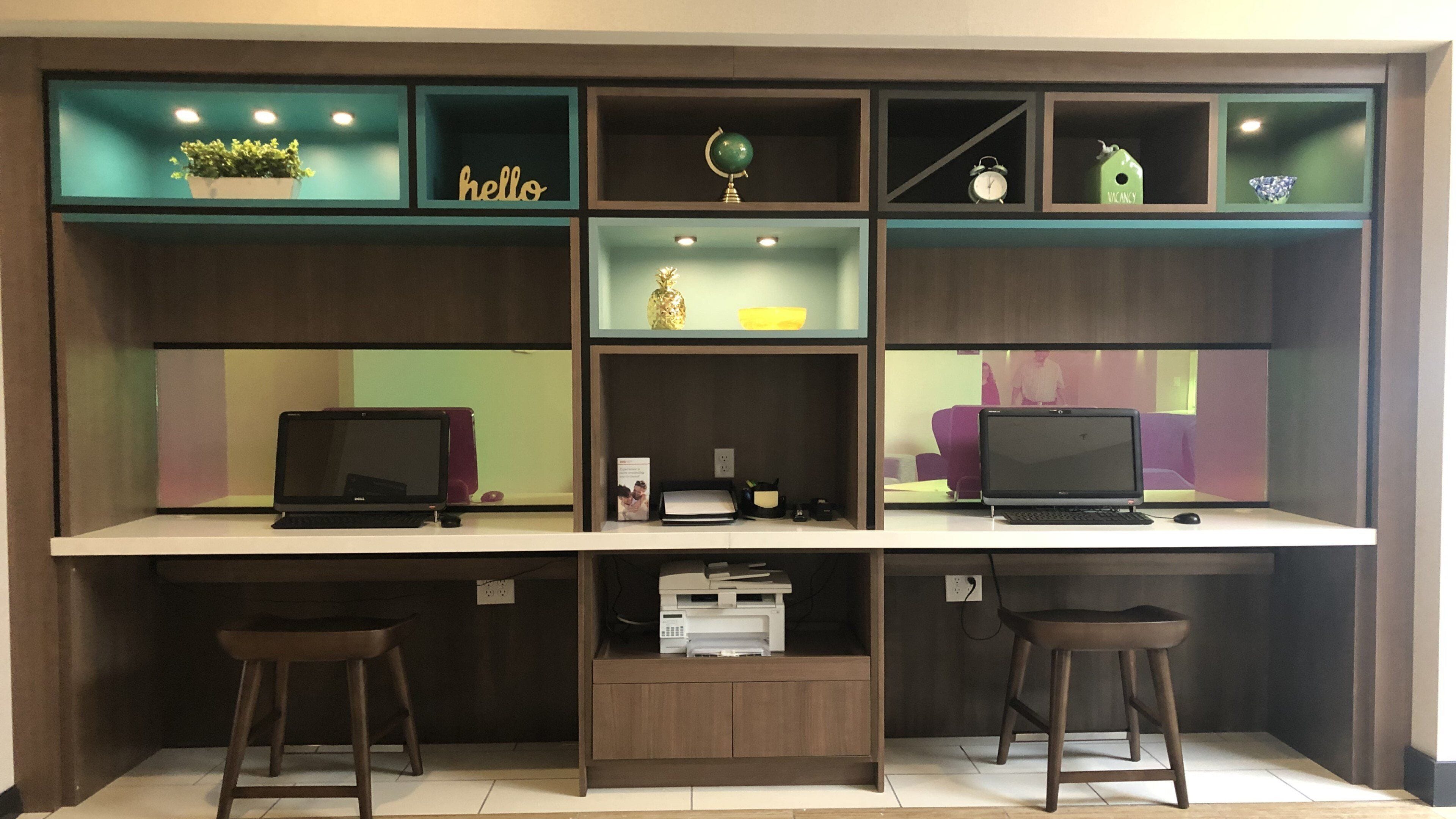 A modern workspace with two desks, each with a laptop, a printer, and a small plant. Shelves above contain decorative items and a globe.