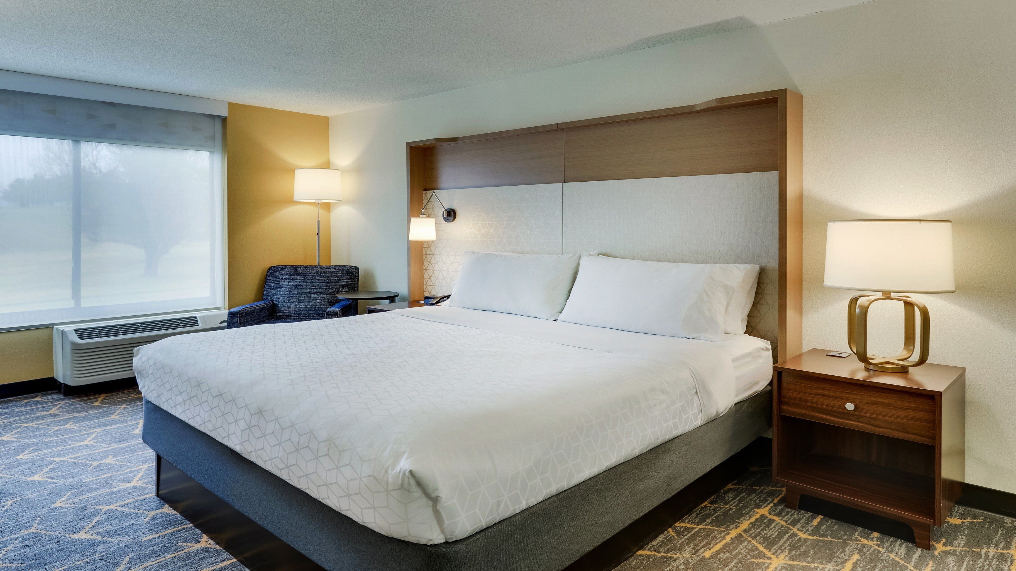 Una habitación de hotel con una cama grande, una silla, una mesita de noche, una lámpara y un calentador.