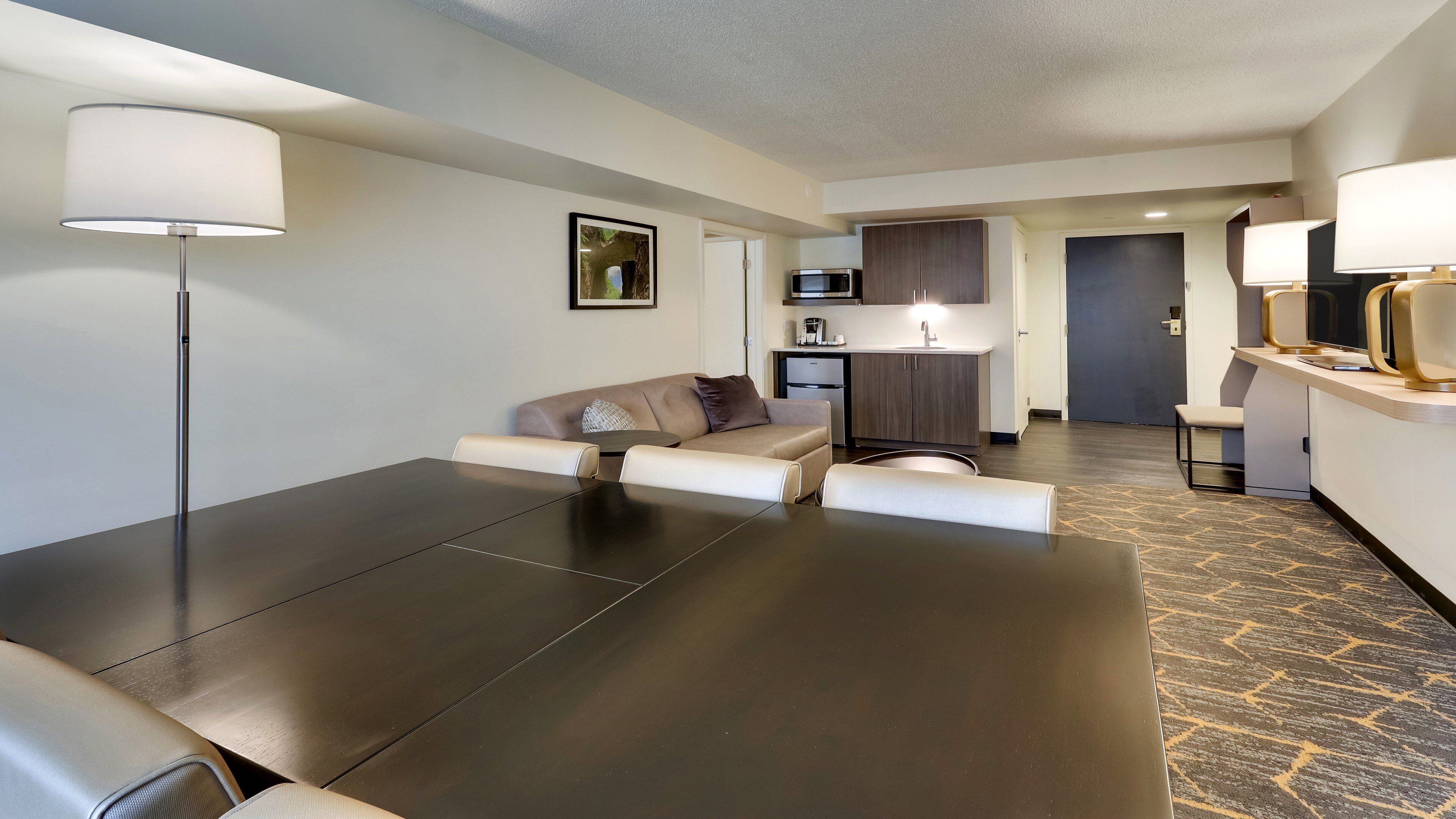 Een moderne hotelkamer met een donker houten tafel, een beige bank, een klein rond tafeltje en een kitchenette op de achtergrond.