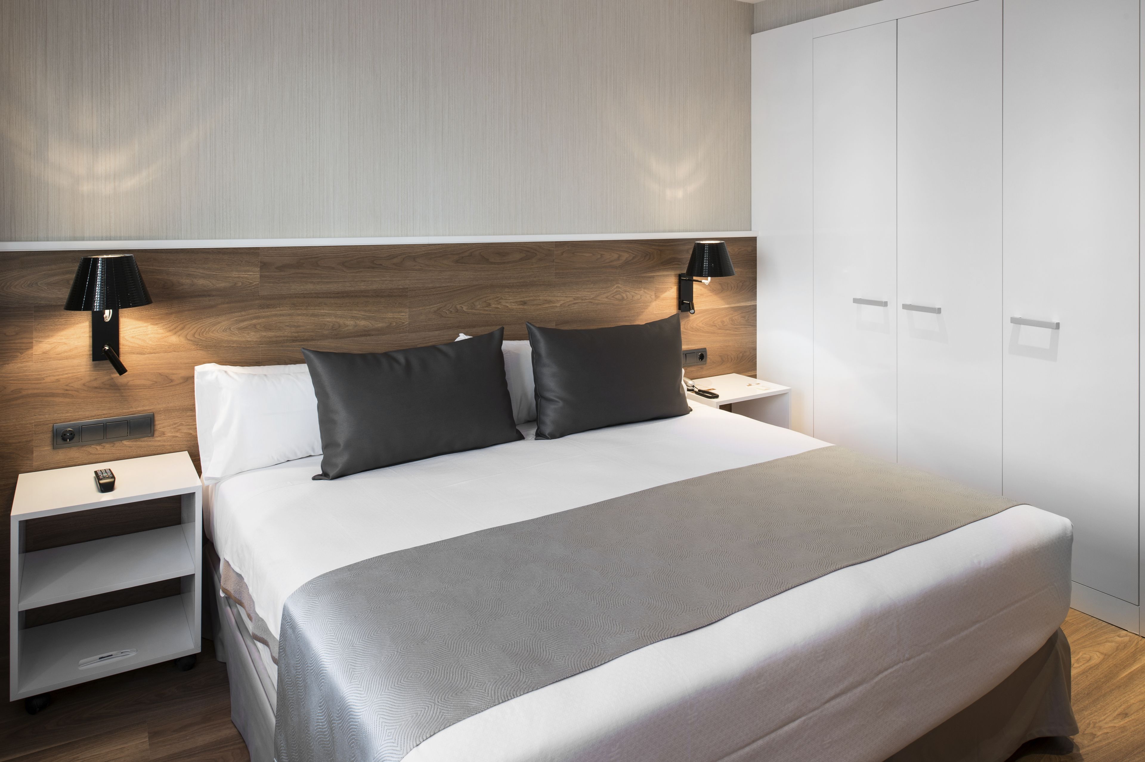 A modern hotel room with a large bed, two bedside tables, a nightstand, a wardrobe, and two wall-mounted lamps.