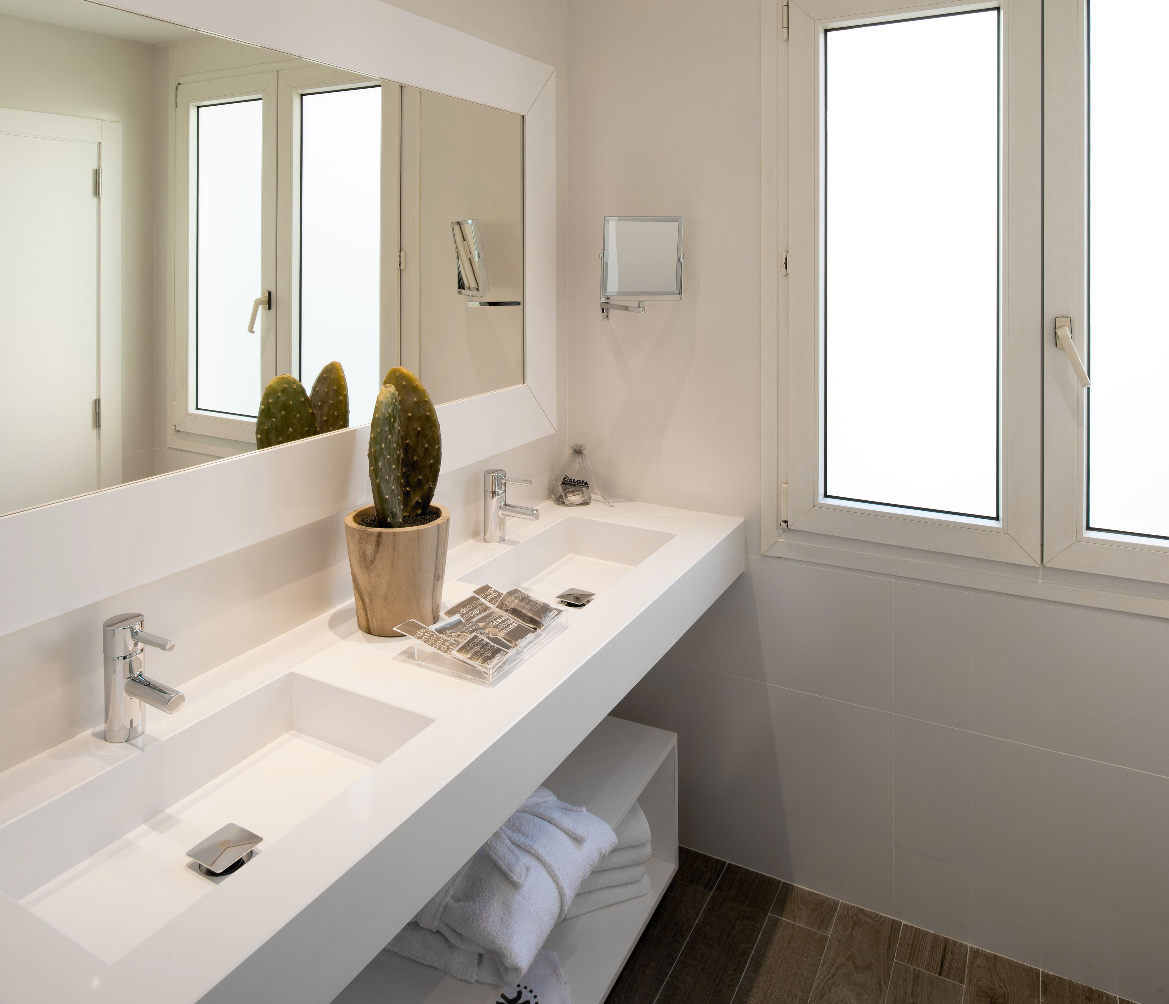 A modern bathroom with a white vanity, a large mirror, a cactus plant, and a window.