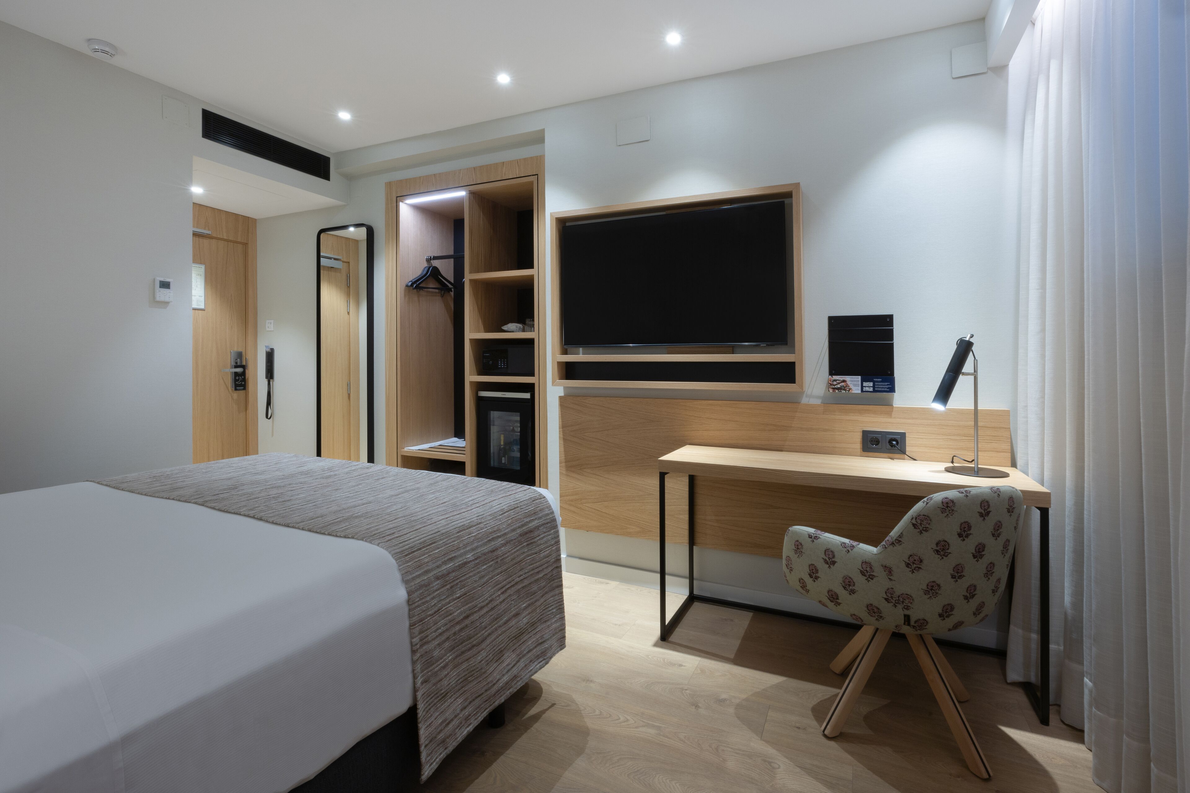 A modern hotel room with a large bed, a desk with a chair, a TV, and a wardrobe.