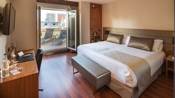 A modern hotel room with a large bed, a desk with a computer, a bench, a TV, and a balcony with chairs. - Catalonia Diagonal Centro (Barcelona)