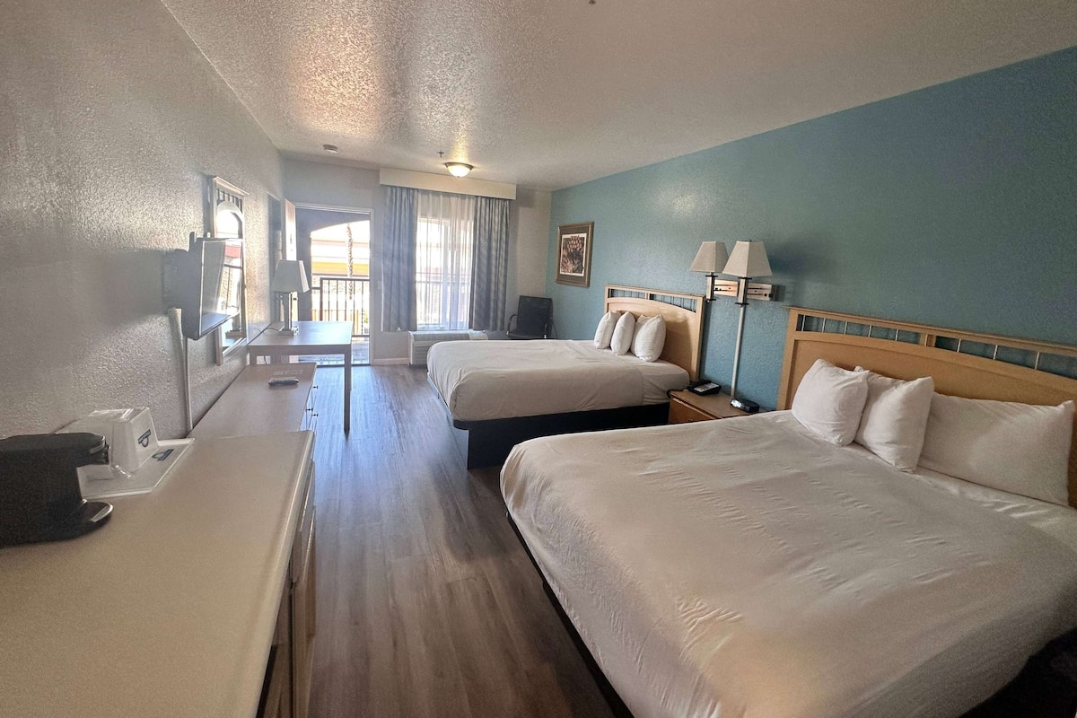 A hotel room with two beds, a desk, and a chair. There is a window with curtains, a lamp, and a painting on the wall.