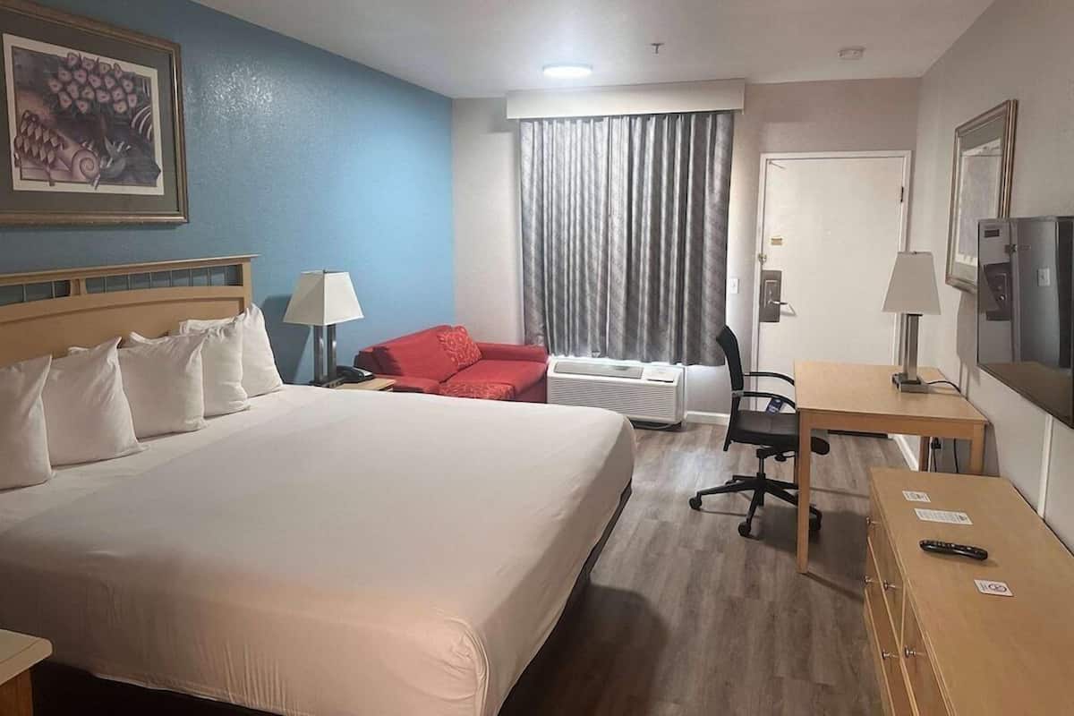 A hotel room with a bed, a desk, a chair, a red sofa, a window with curtains, and a painting on the wall.