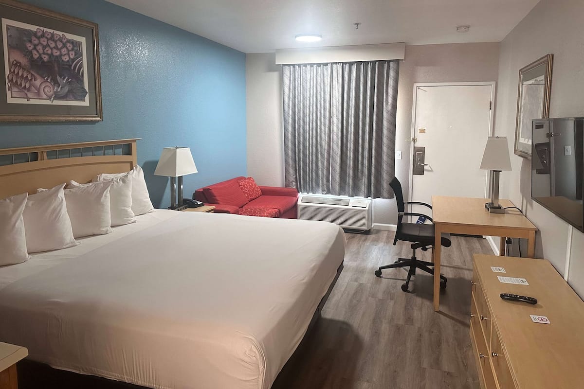 A hotel room with a large bed, a desk, a chair, a red sofa, a window with curtains, and a refrigerator.