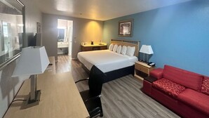 Habitación de hotel con cama, escritorio con lámpara, sofá rojo, mesita de noche y un cuadro en la pared.