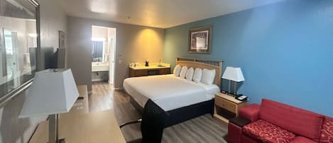 A hotel room with a bed, a desk with a lamp, a red sofa, a nightstand, and a wall-mounted picture.