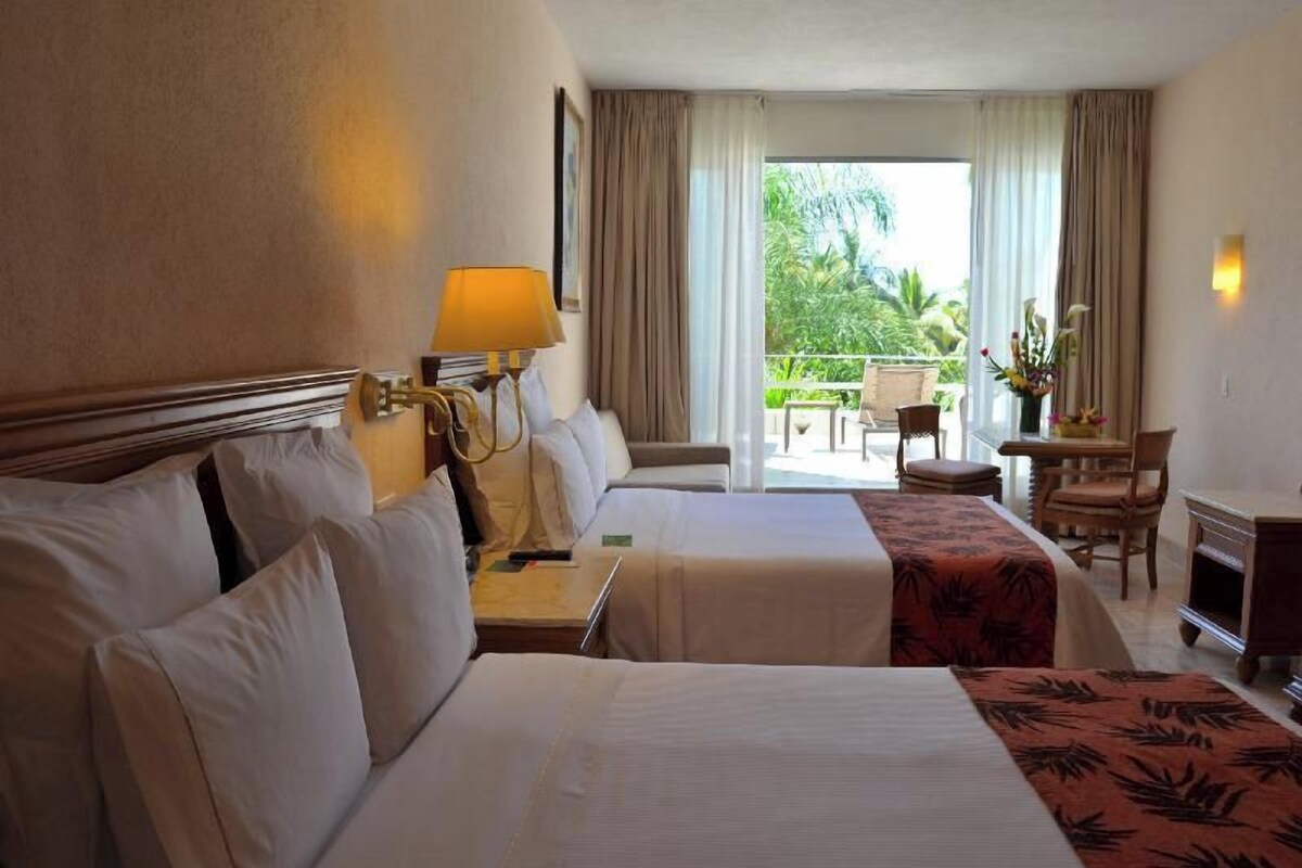 Une chambre d’hôtel avec deux lits, un bureau, une chaise, une petite table et un balcon donnant sur un espace verdoyant.