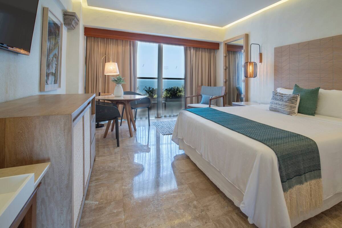 A hotel room with a large bed, a desk, a chair, a small table, and a balcony with curtains.