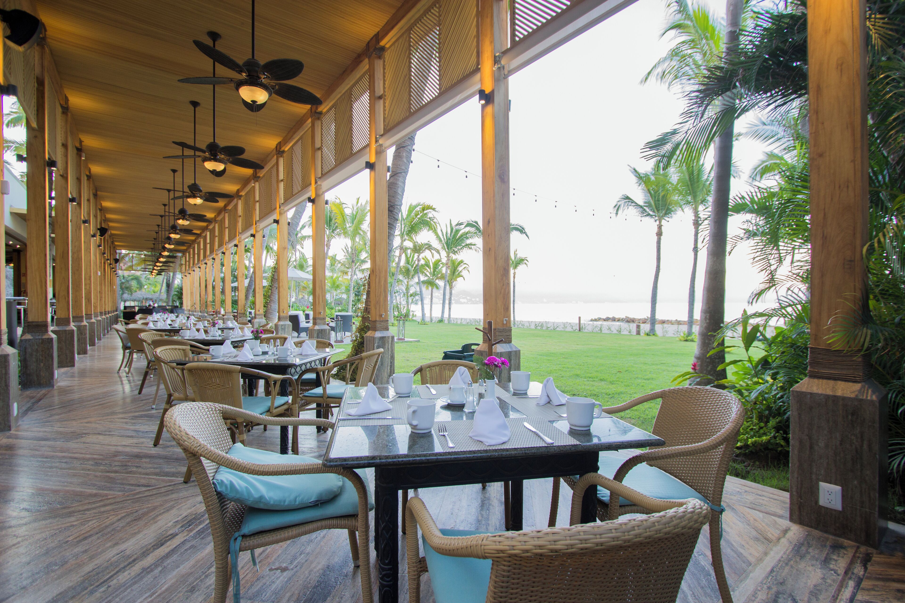 A restaurant with outdoor seating overlooking a beach, featuring wicker chairs and a long table set for dining.