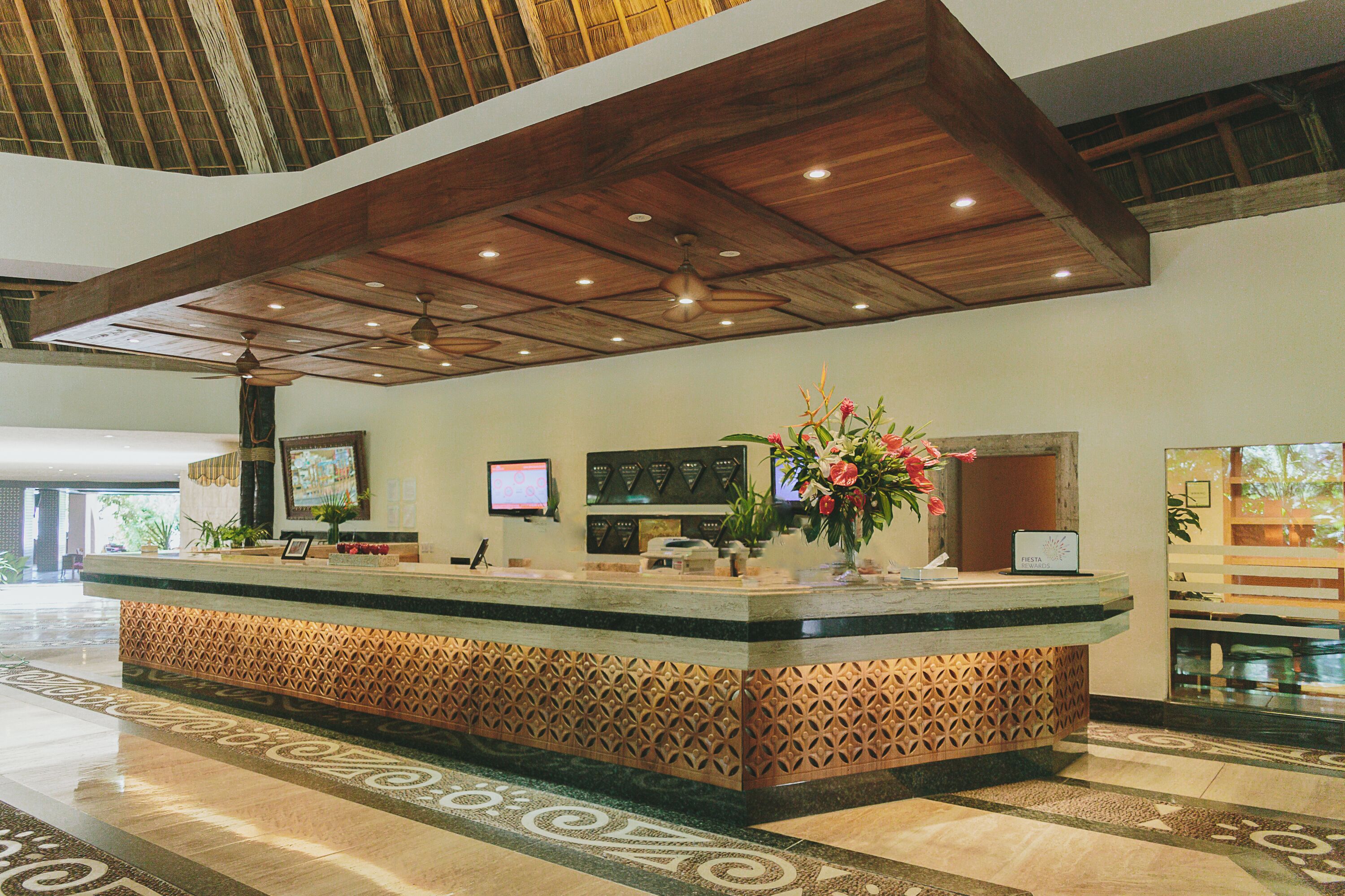 Un área de recepción con techo de madera, una mesa de recepción, un monitor, una planta y un panel decorativo en la pared.