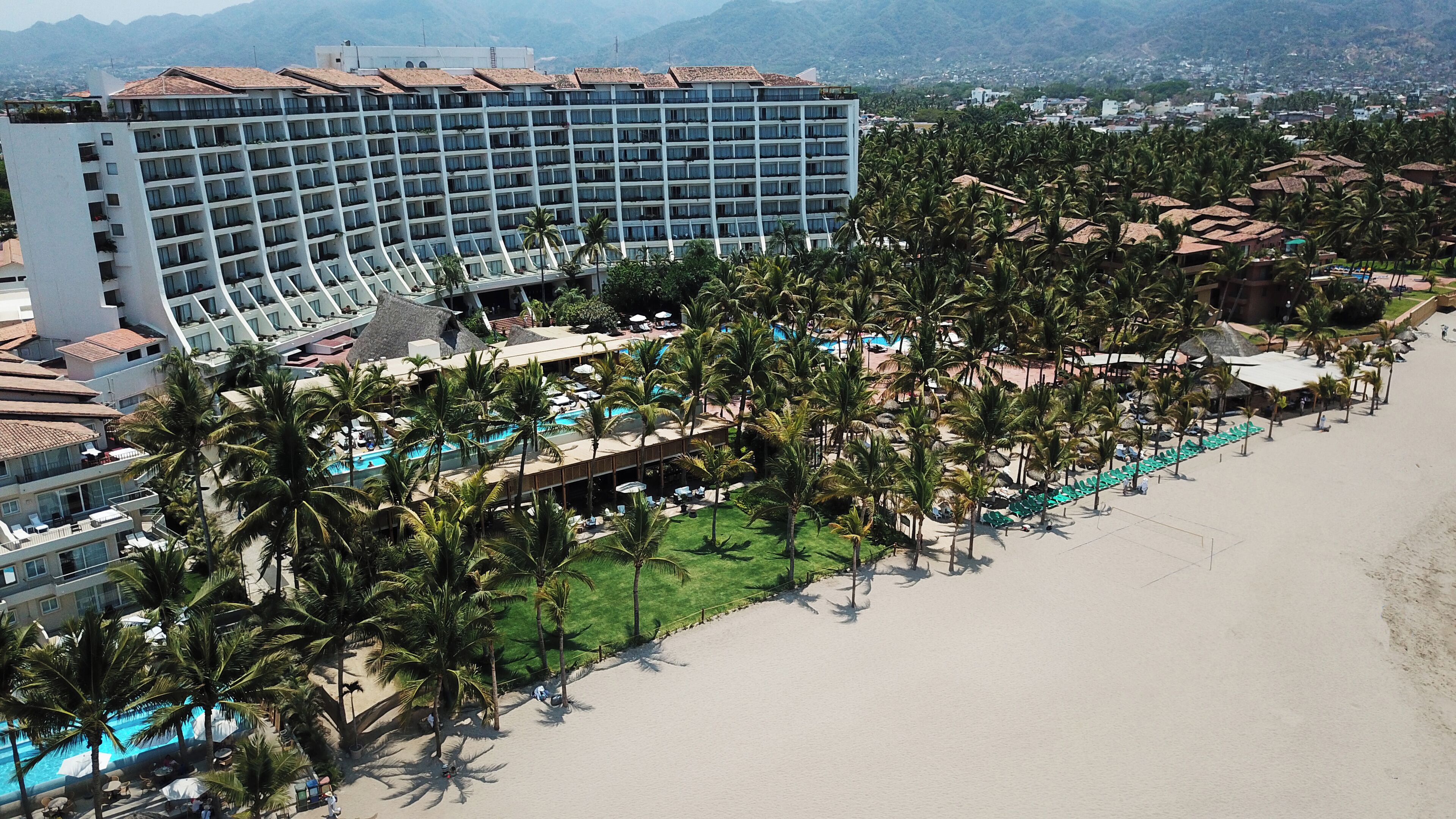 Um resort com um edifício grande, piscina e área de praia com palmeiras.