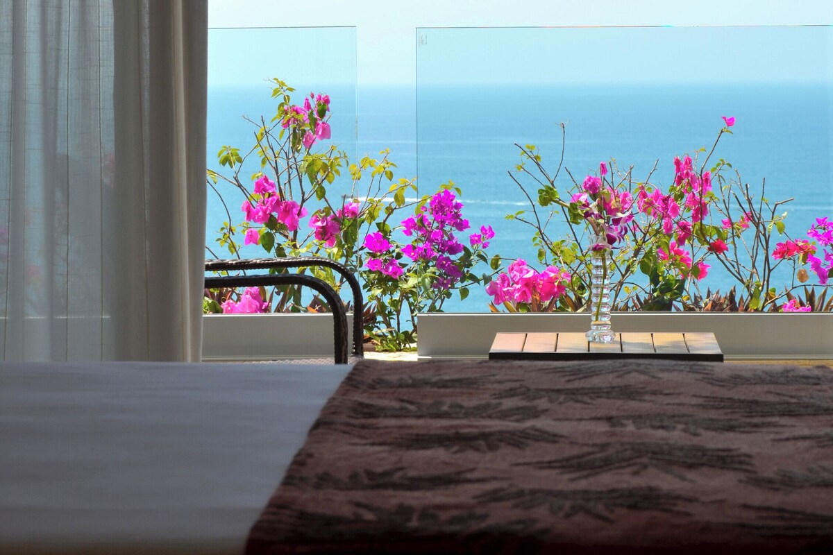 A balcony with a table, pink flowers, and a view of the sea and a parachute.