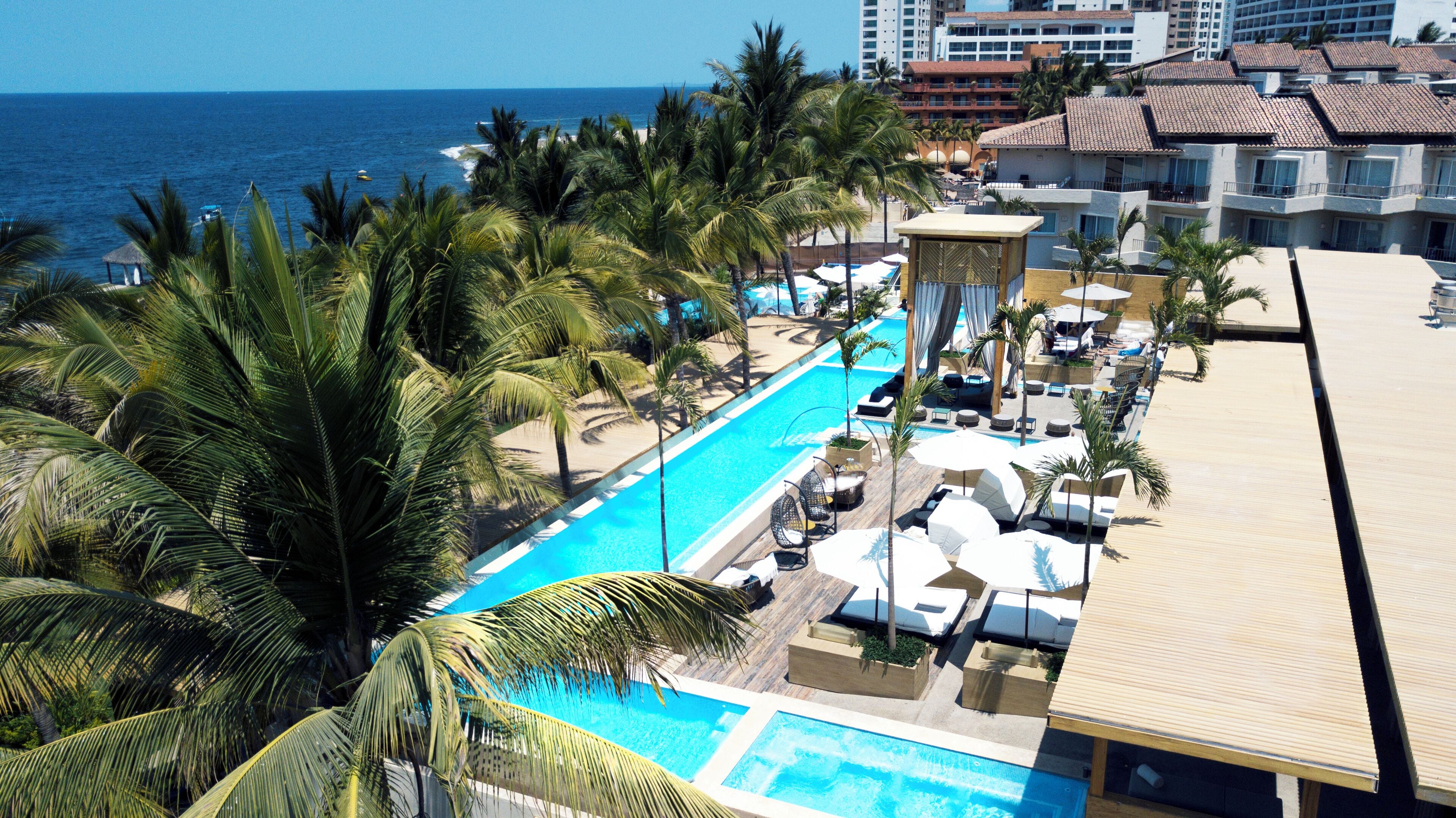 Zona junto a la piscina con sillones de descanso y sombrillas, rodeada de palmeras y vista al océano.