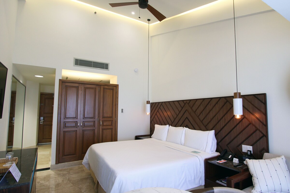 A hotel room with a large bed, a desk with a computer, a mirror, a wardrobe, and a ceiling fan.