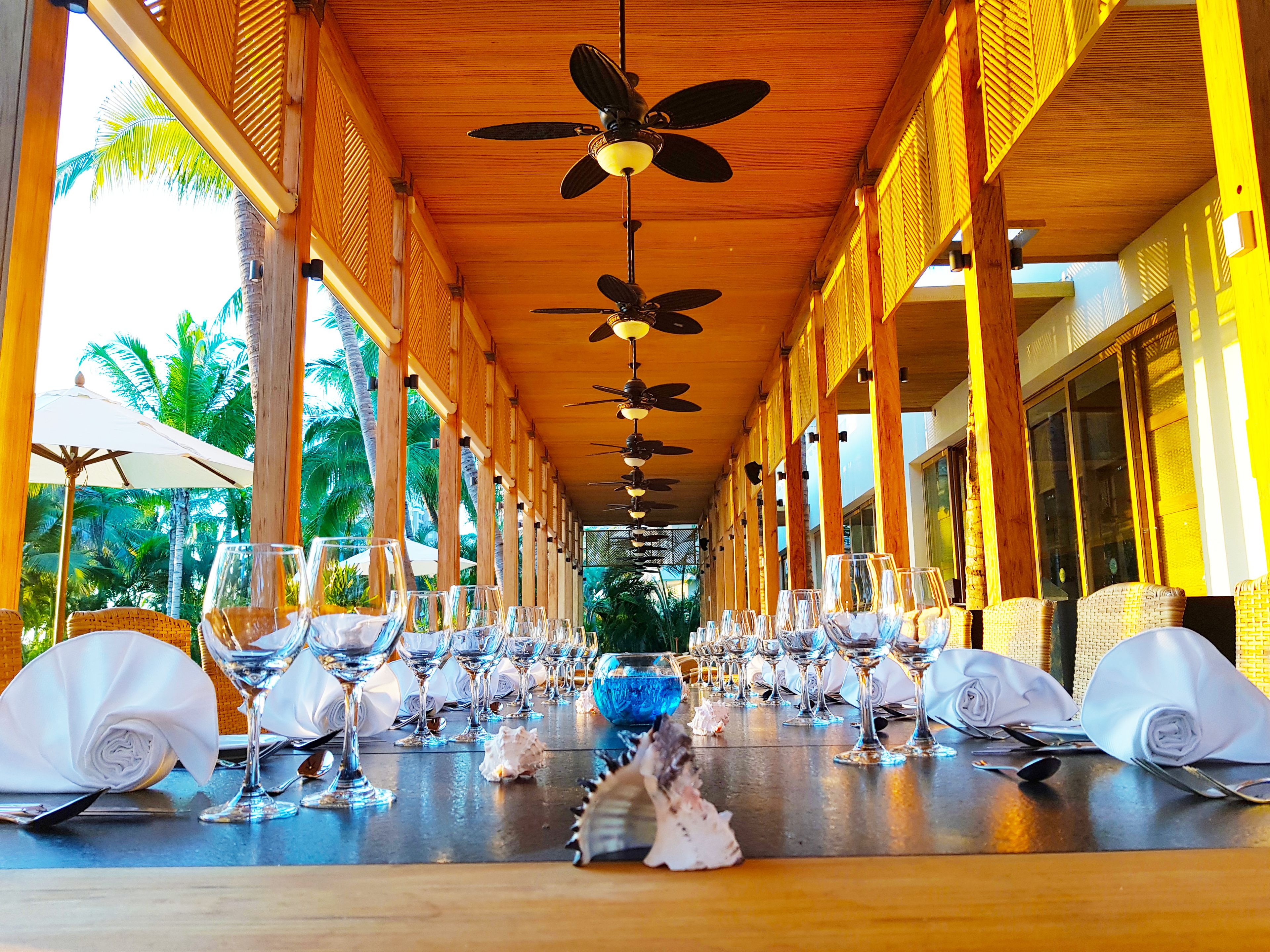 Un espacio de comedor al aire libre techado, con varias mesas dispuestas para comer, ventiladores de techo y palmeras al fondo.