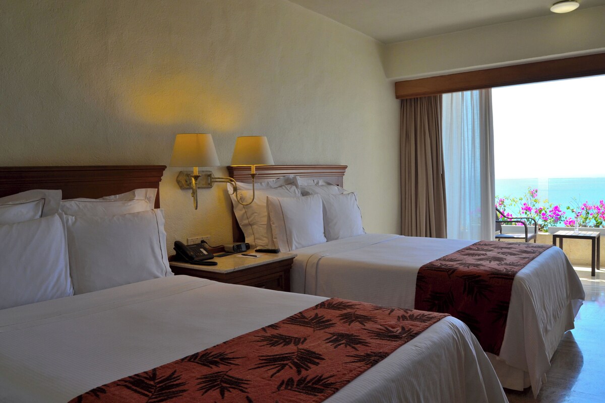 A hotel room with two beds, a nightstand, a lamp, and a view of the ocean.
