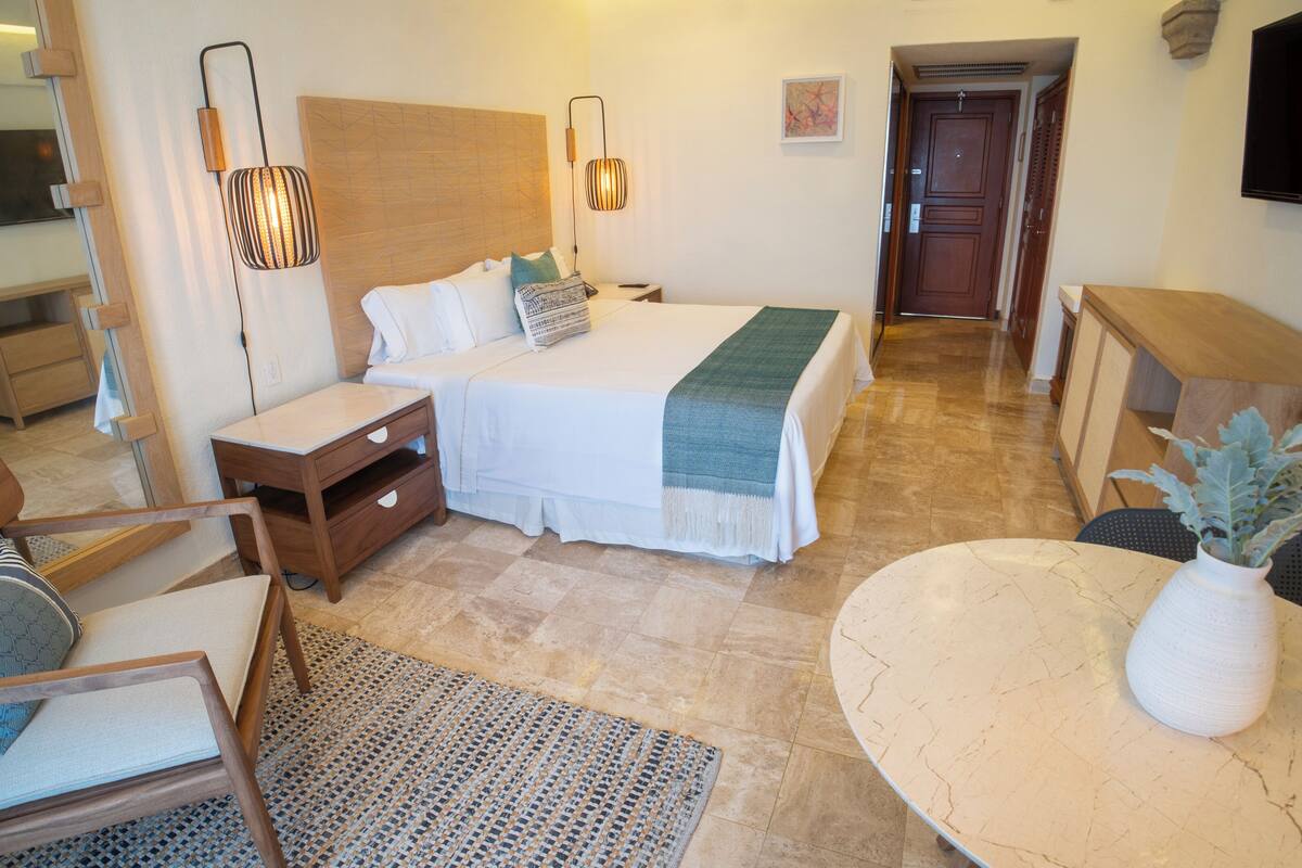 A hotel room with a bed, bedside tables, a chair, a small table, and a TV mounted on the wall.