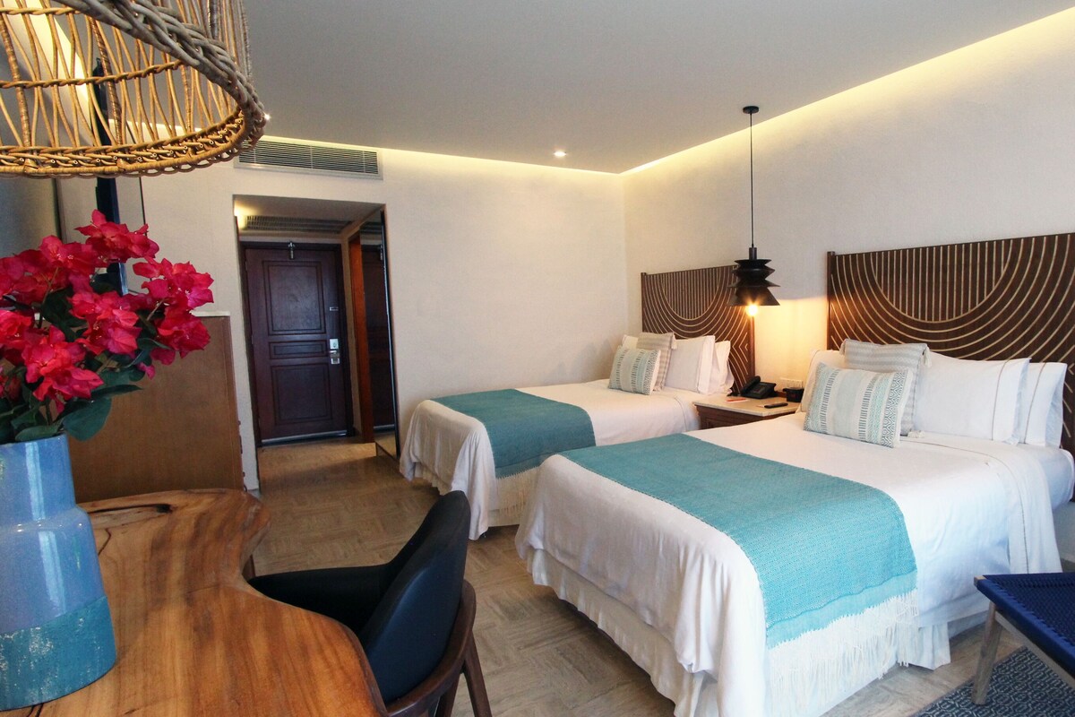 A hotel room with two beds, a desk, a chair, a mirror, and a wicker chair.
