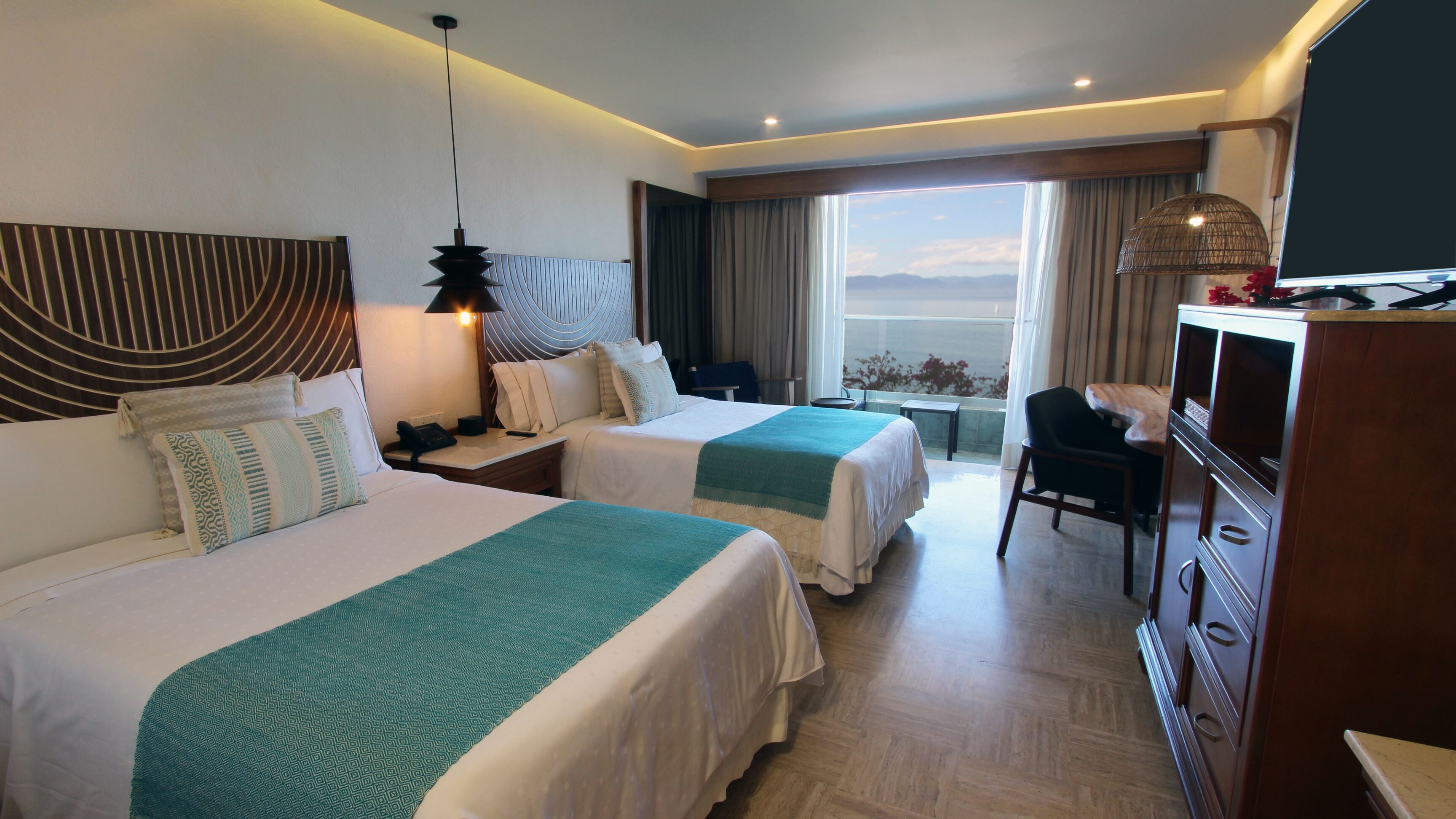 Quarto de hotel com duas camas, uma escrivaninha, uma cadeira, uma televisão e vista para o mar.