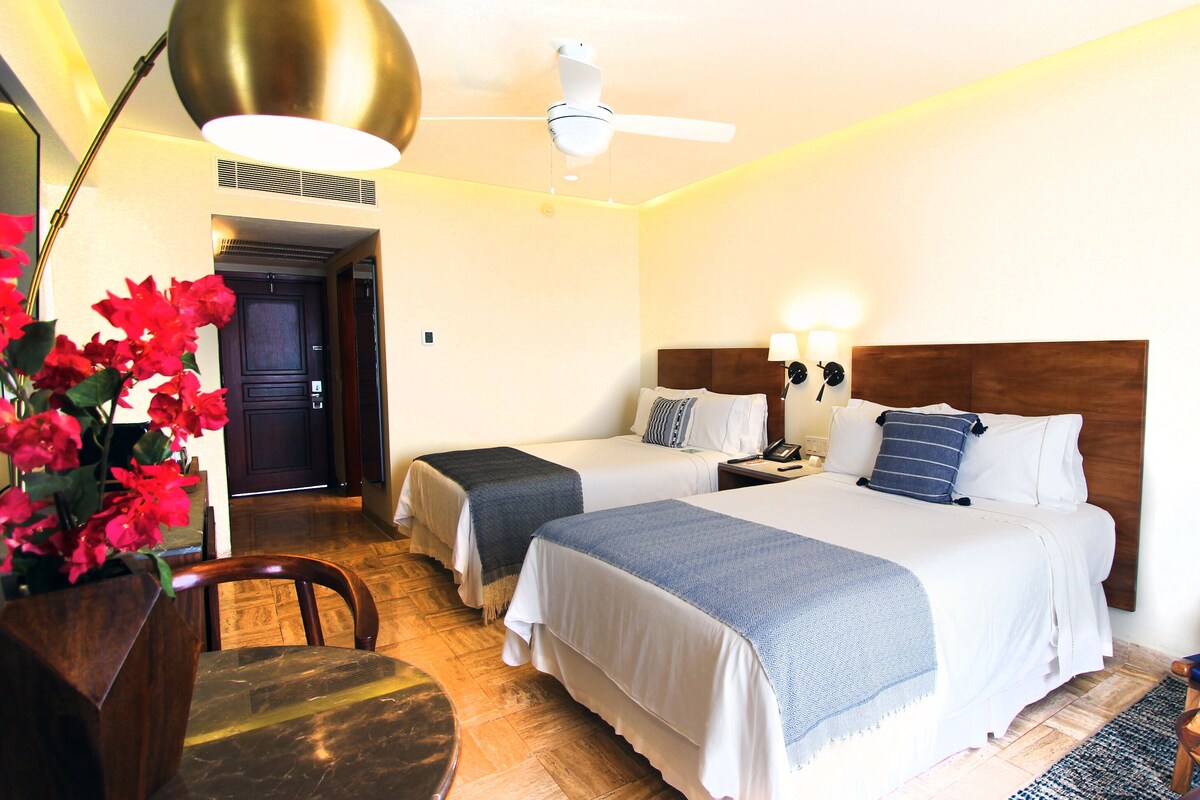 A hotel room with two beds, a desk, a chair, a ceiling fan, and a wall-mounted lamp.