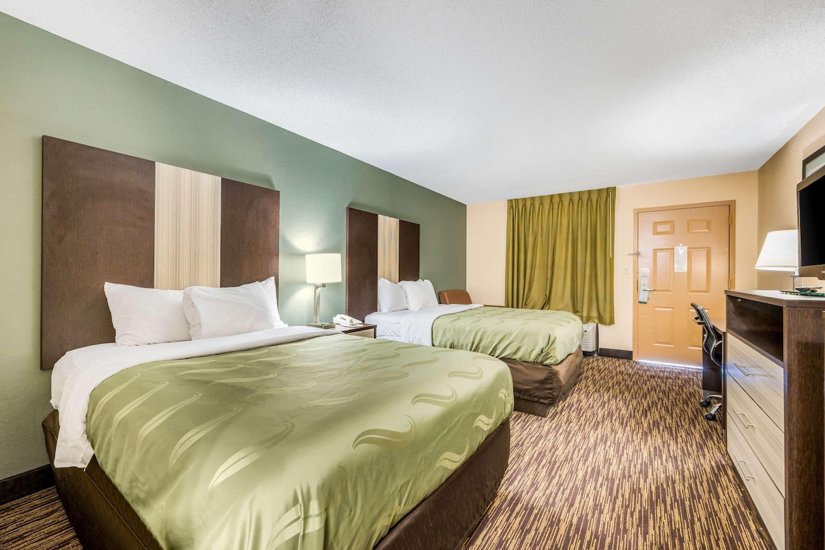 A hotel room with two beds, a desk with a lamp, a television, and a door.