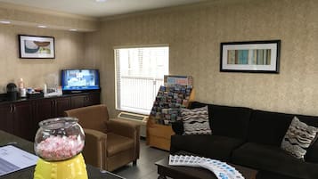 A living room with a black sofa, a brown armchair, a coffee table with magazines, a TV on a stand, a bookshelf with books, and a window with blinds.
