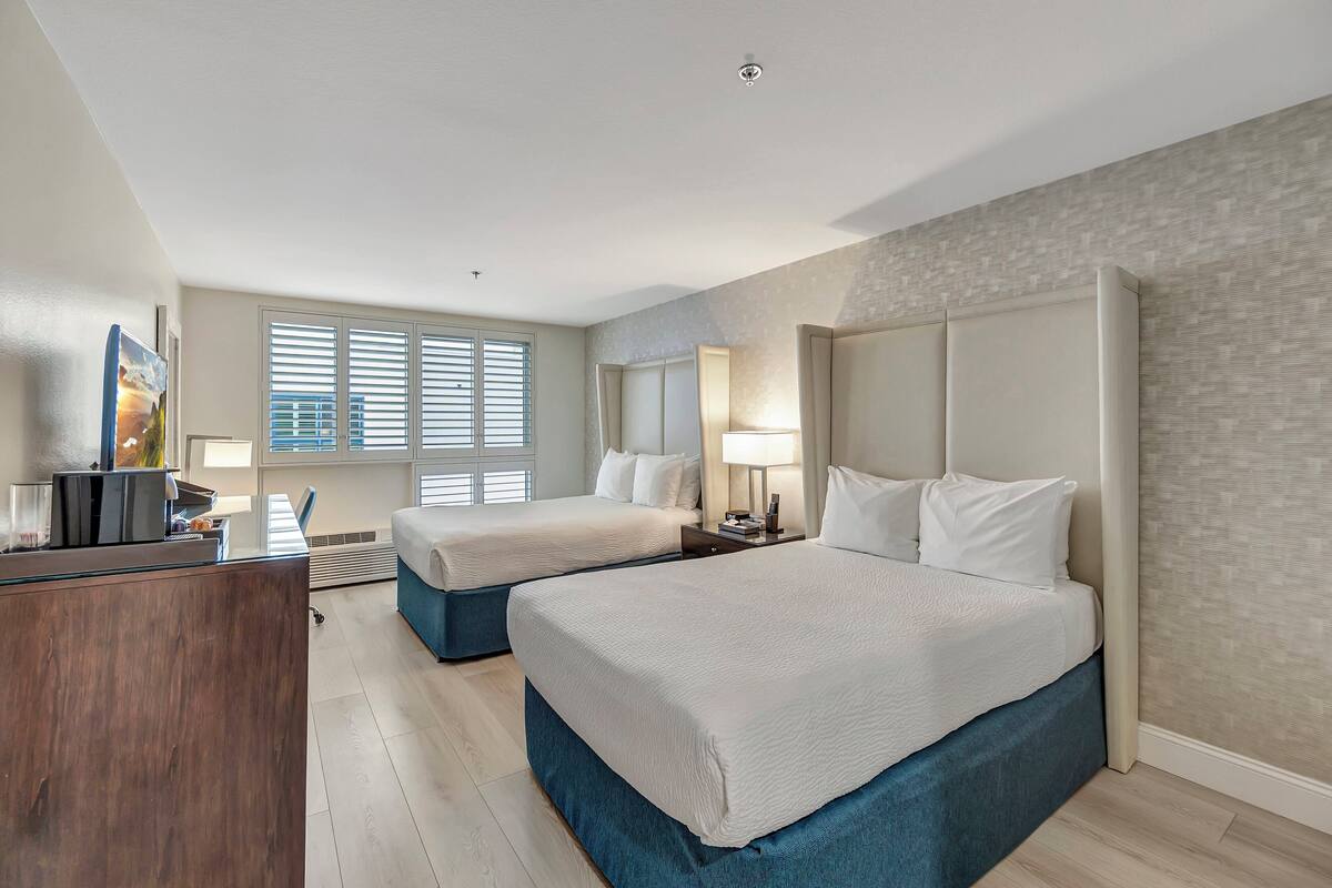 A hotel room with two beds, a desk, and a window with blinds.