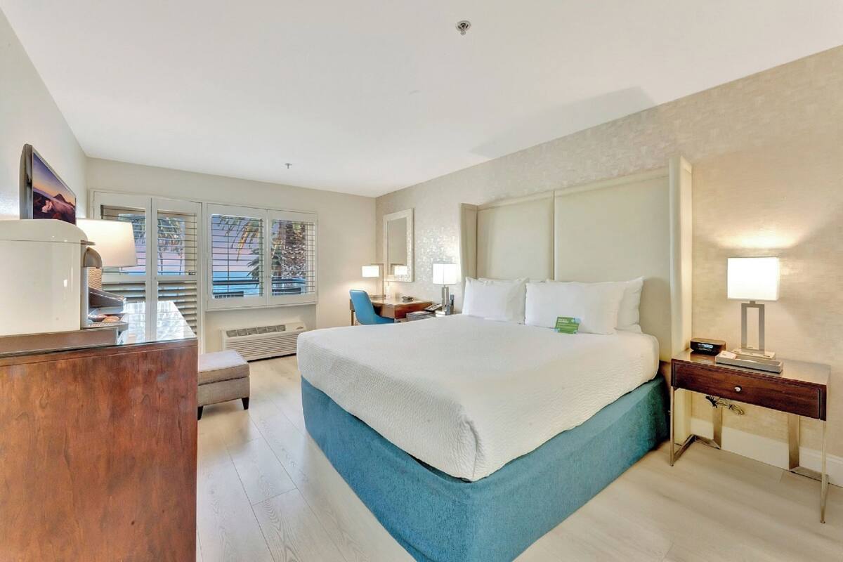 A hotel room with a large bed, a desk, a chair, and a window with blinds.