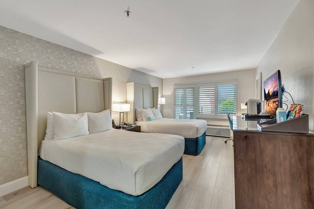A hotel room with two beds, a desk, a TV, and a window with blinds.