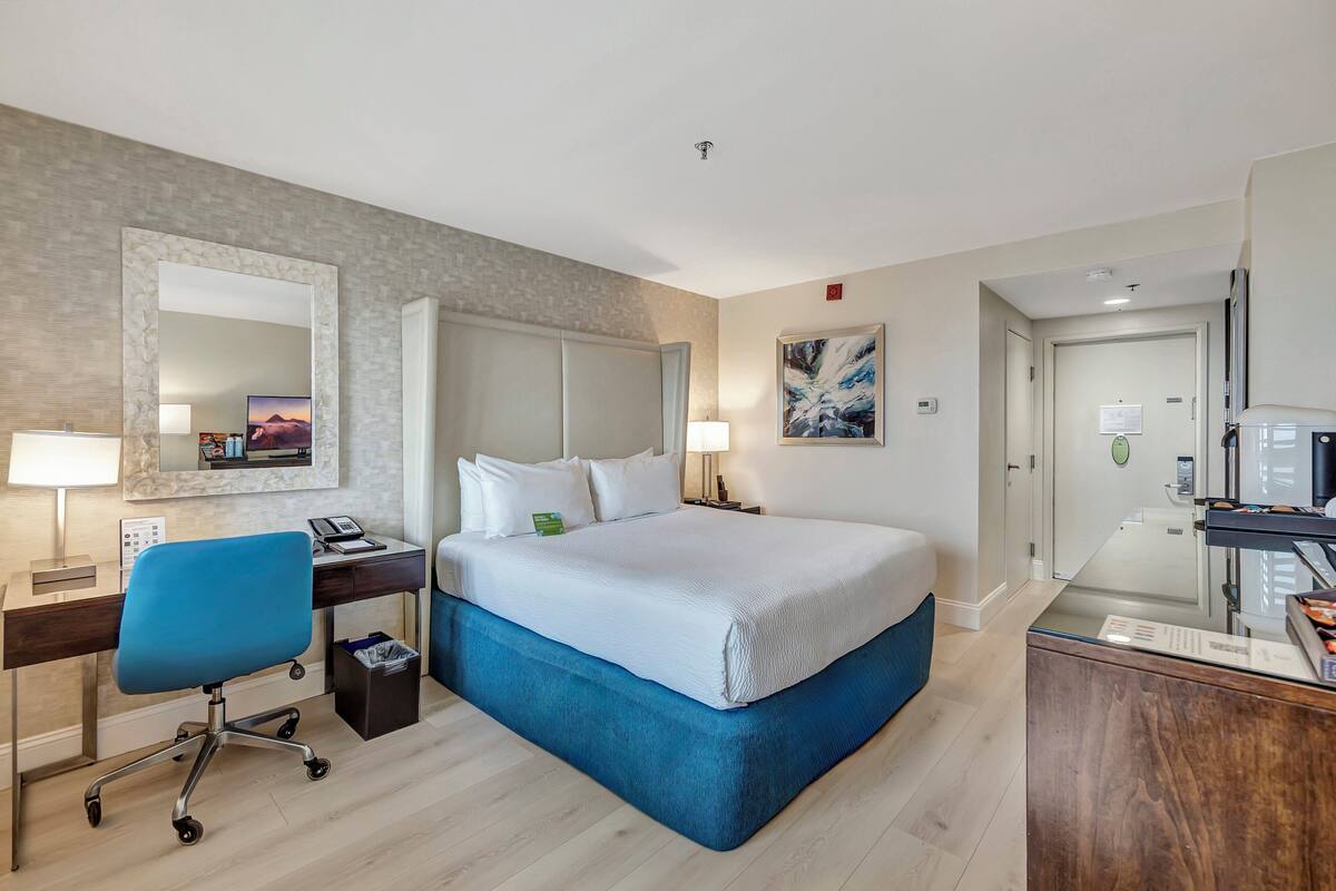 A hotel room with a large bed, a desk with a chair, a mirror, and a painting on the wall.