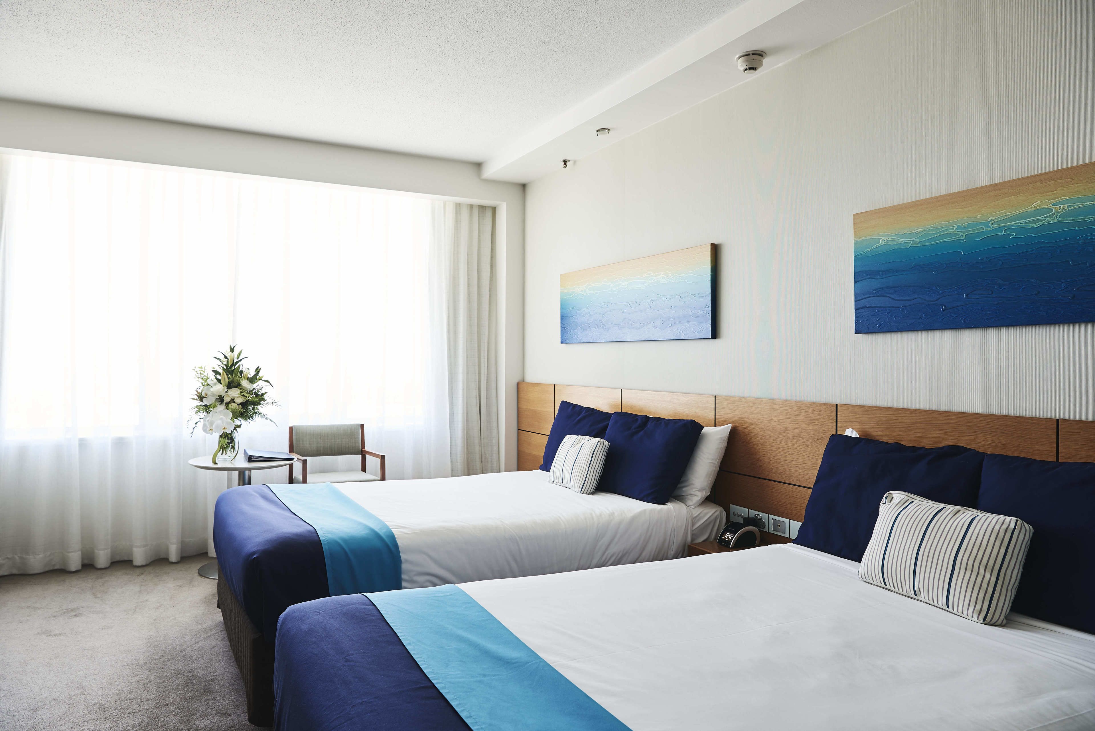 Habitación de hotel con dos camas, una mesita con un jarrón de flores y vistas al mar a través de amplios ventanales.