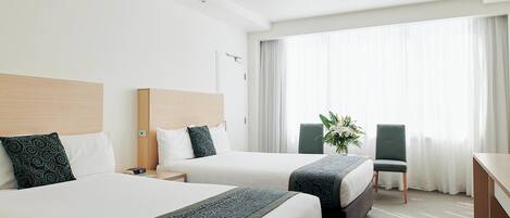 A hotel room with two beds, a desk, a chair, and a large window with curtains.