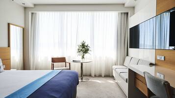 A hotel room with a bed, a desk, a chair, a sofa, a TV, and a window with curtains.