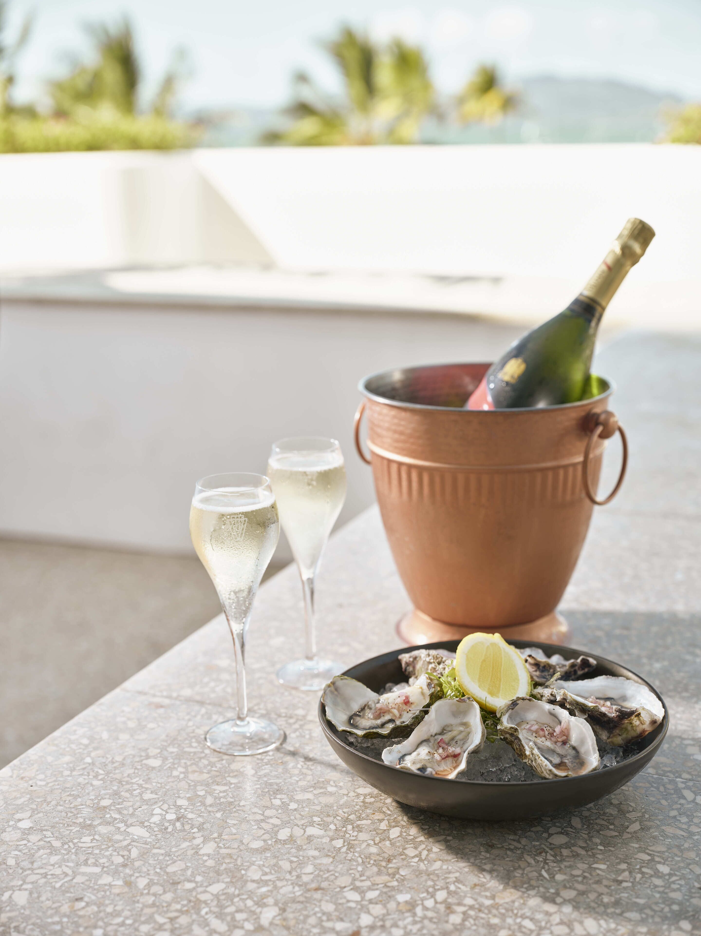 A table with a copper bucket containing a bottle of champagne, two glasses of champagne, and a plate of oysters garnished with lemon.